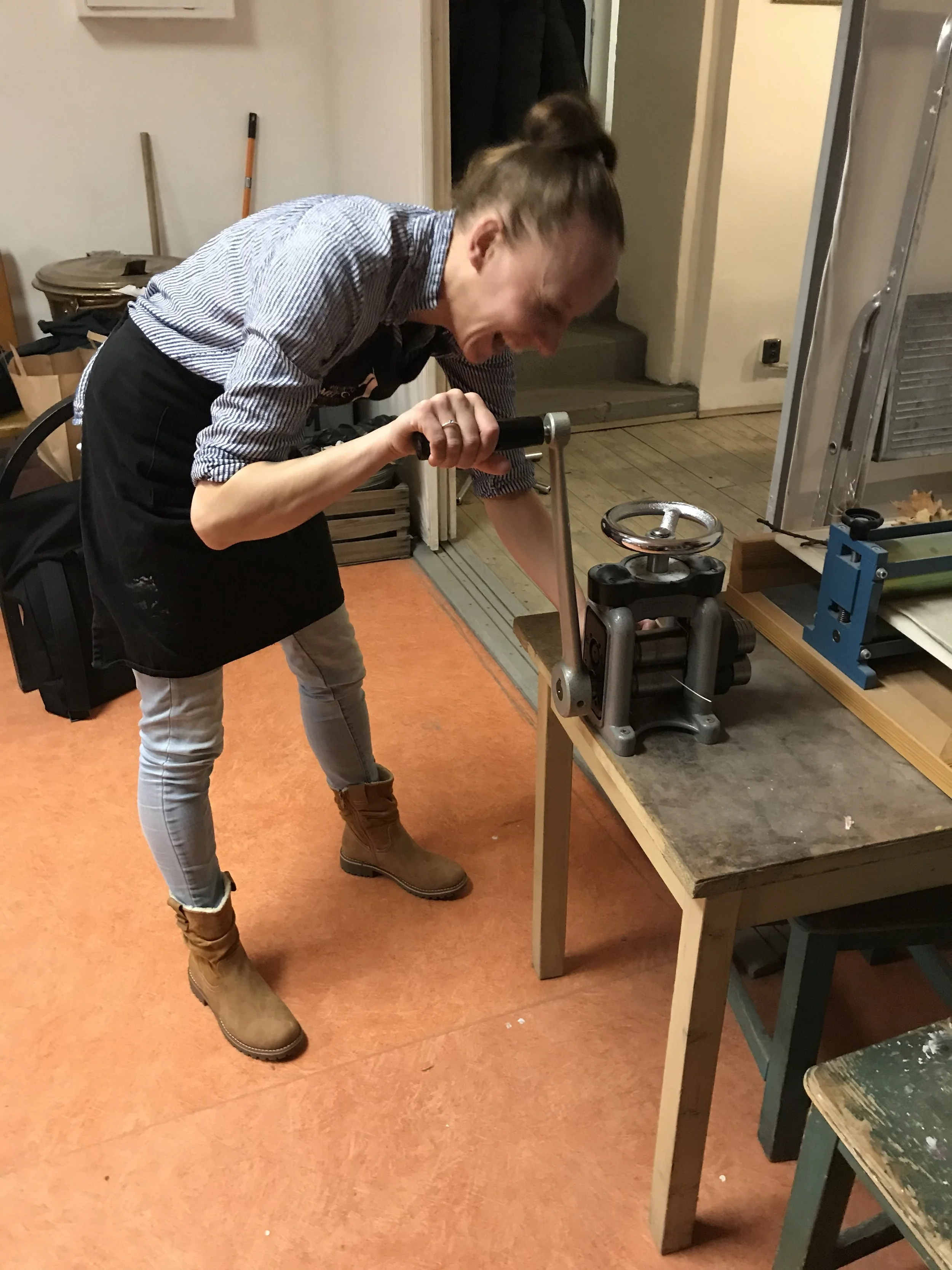 A person with their hair tied back, wearing a striped shirt, dark apron, beige pants, and brown boots, is leaning over and operating a manual pasta machine on a wooden worktable in a room with orange flooring and various tools and furniture around.