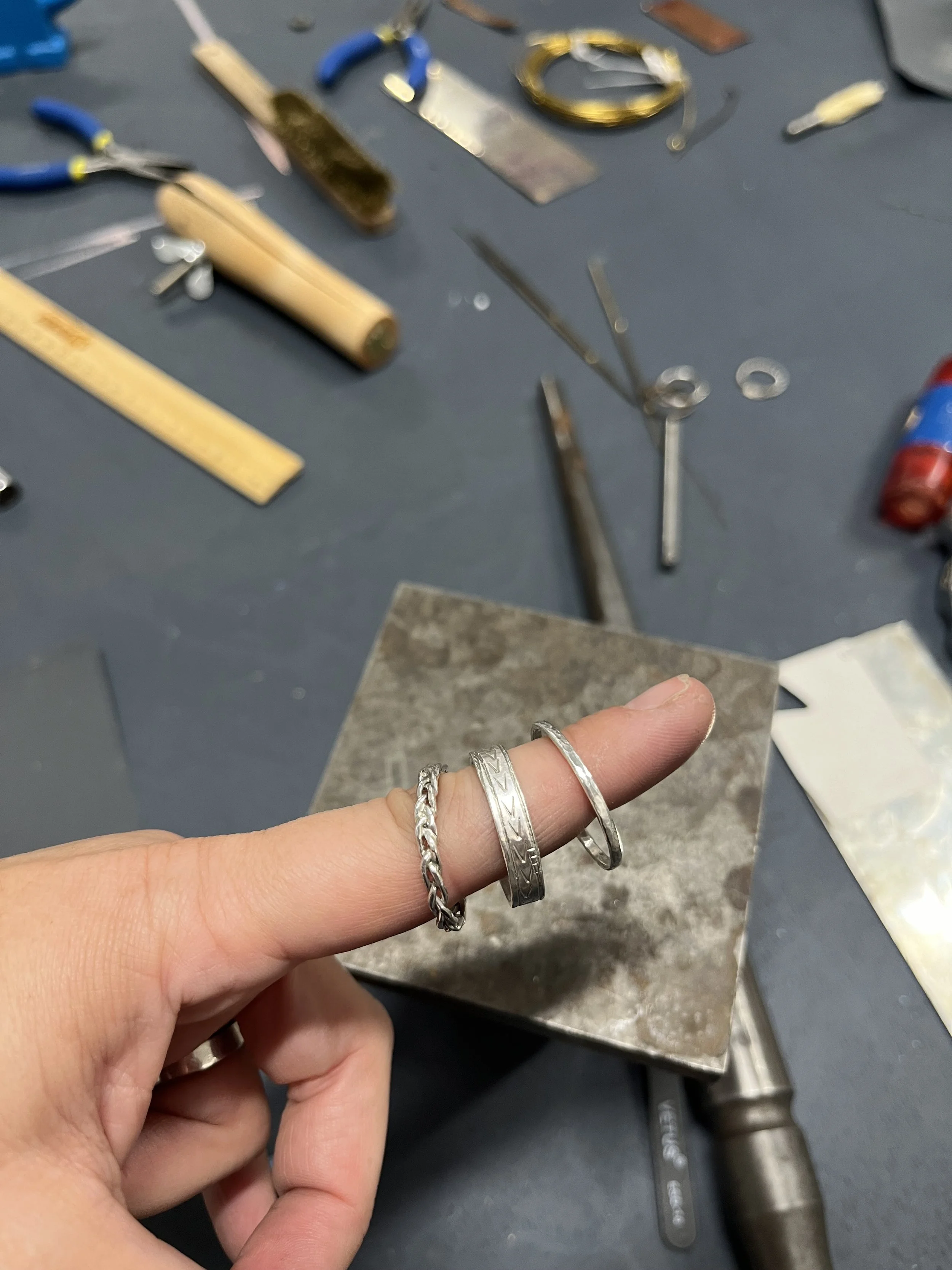A person holding three silver rings with various patterns and designs, with jewelry-making tools and supplies on a workbench in the background.