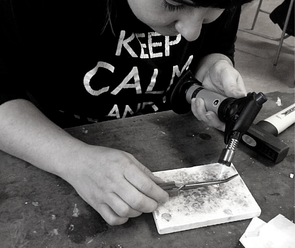 A person operating a heat gun and using tweezers to work on a small piece of material on a workbench.