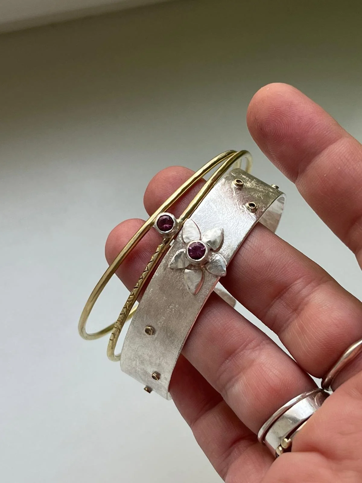 Close-up of a hand holding jewelry, including a silver bracelet with a flower and purple gemstone, a gold bangle, and a gold ring, against a plain background.