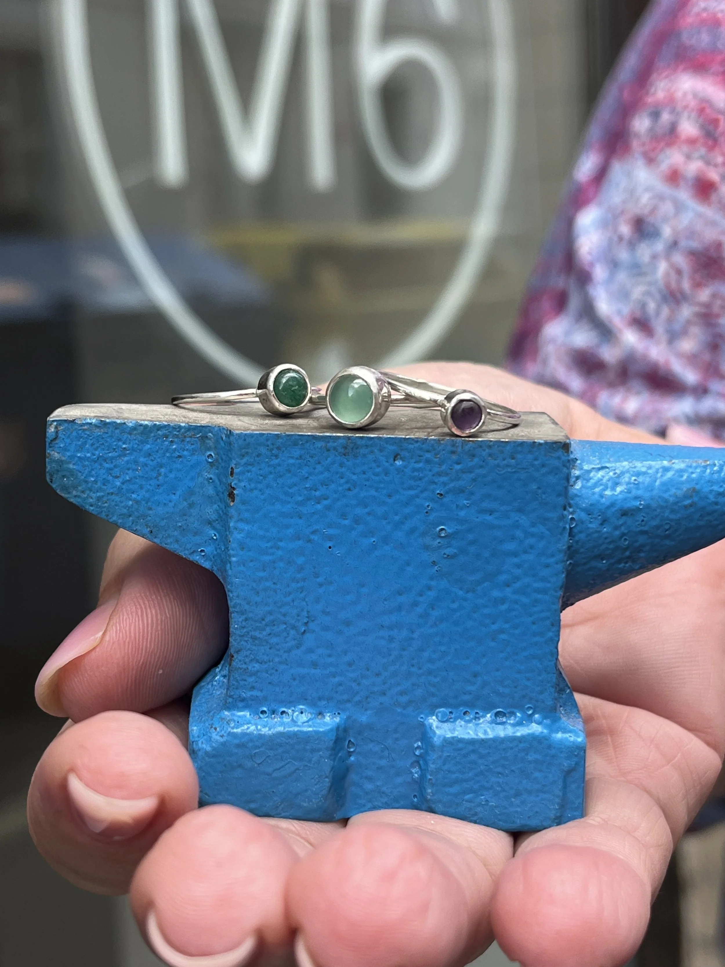 Close-up of a hand holding a blue object with three silver rings with colored stones, with a blurry background showing a neon sign.