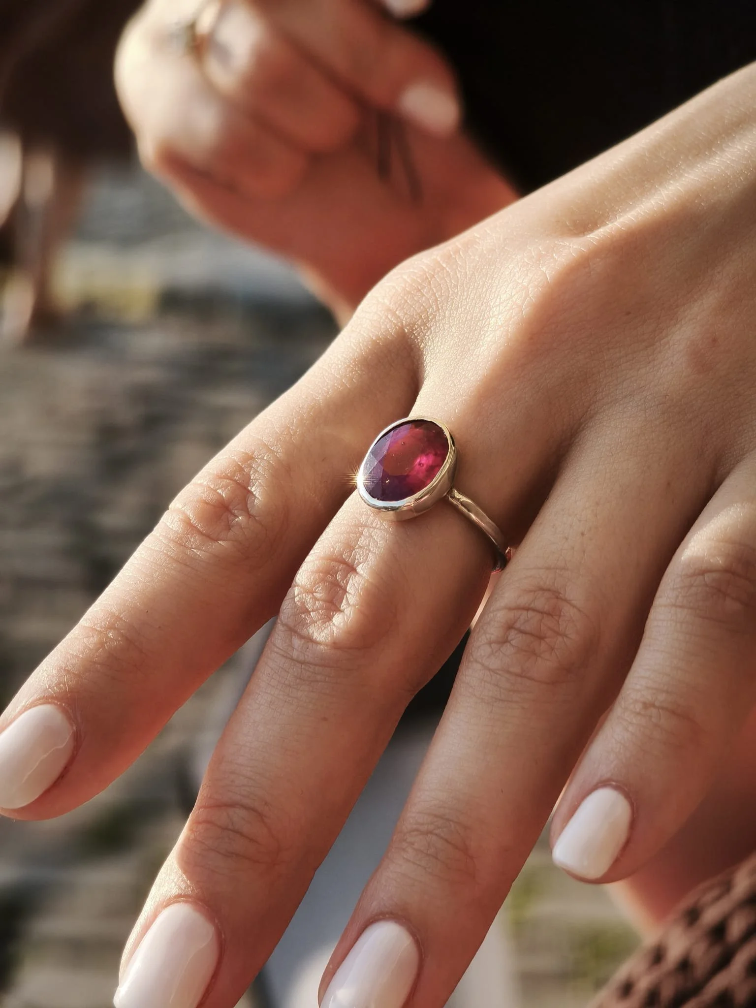 A hand with a ring featuring a large oval-shaped pink gemstone, with a person's face in the background out of focus.