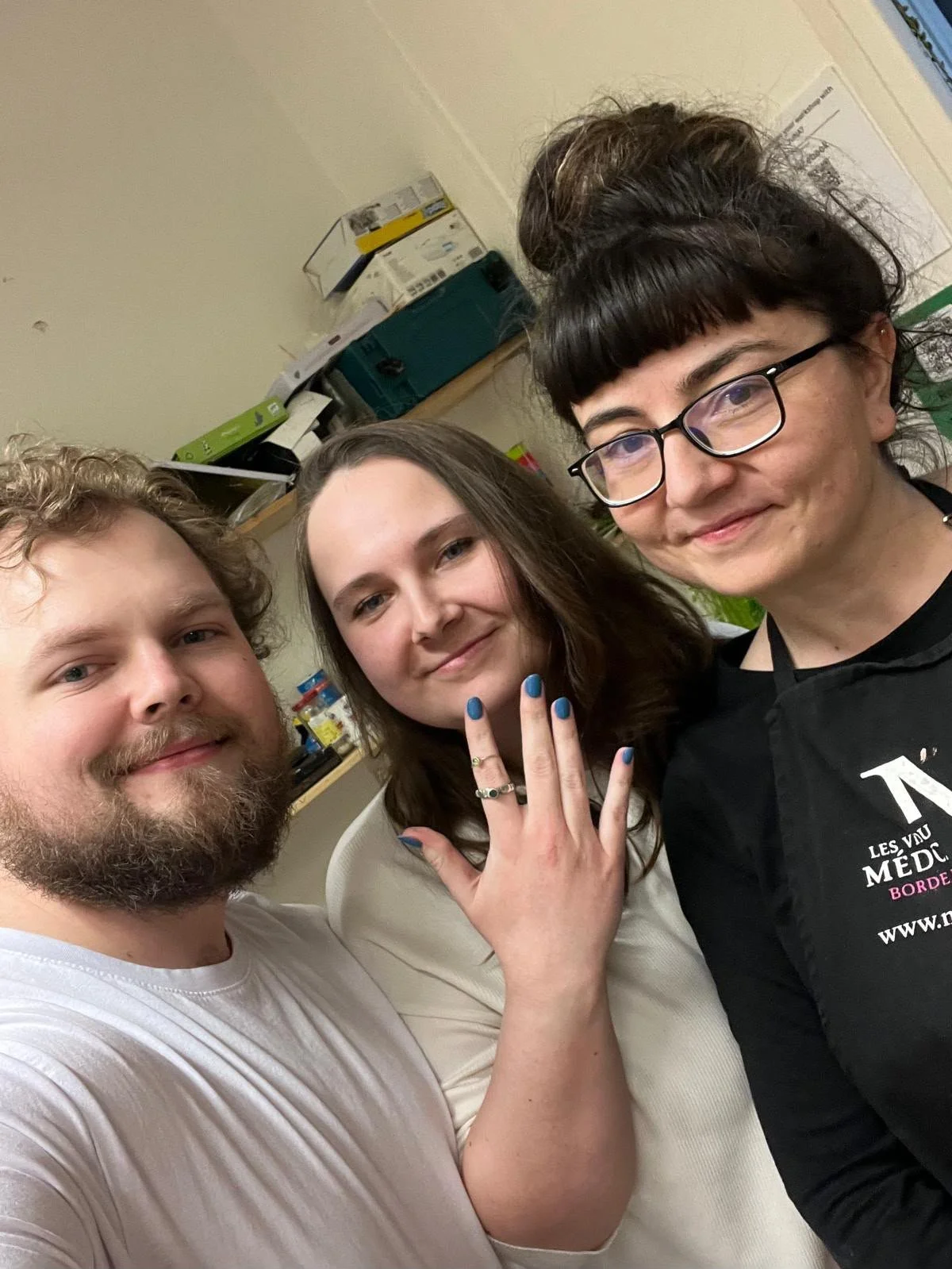 Three people taking a selfie in a room, smiling, with a woman showing her ring and nails.
