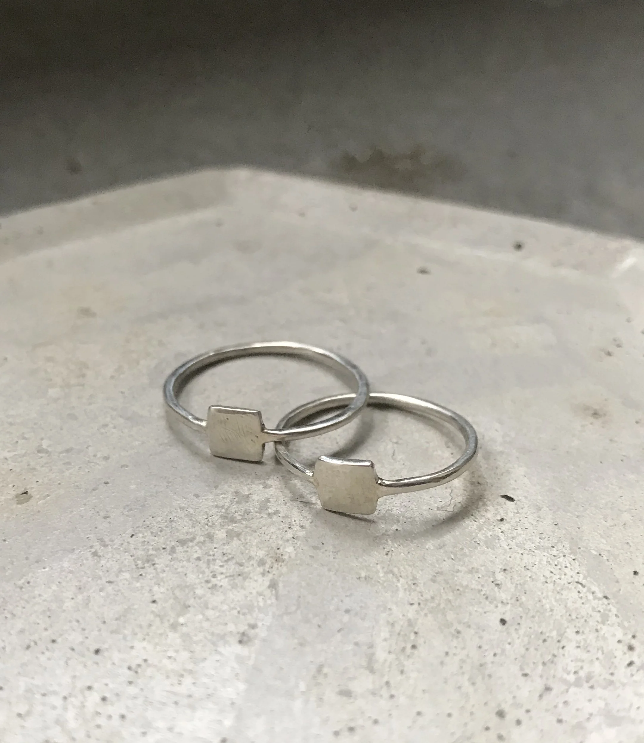 Close-up of two silver rings with heart-shaped charms on a light gray textured surface, with part of a silver object visible above.