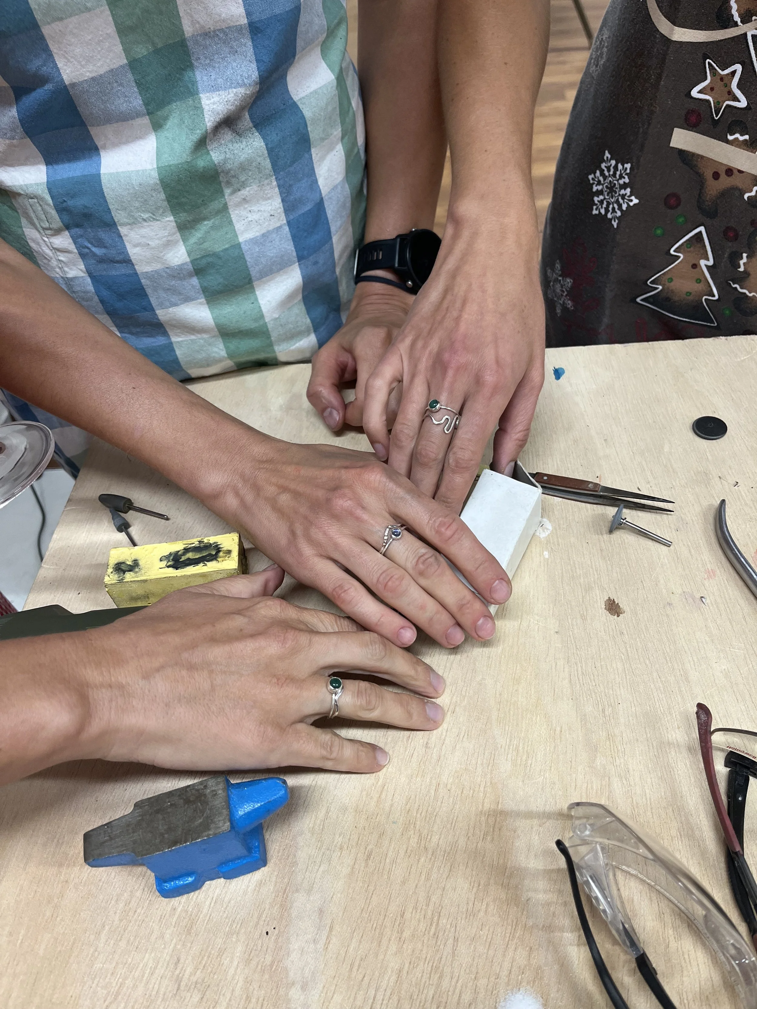 Four hands with rings and watches working together on a jewelry or craft project at a workbench, with tools and materials scattered around.