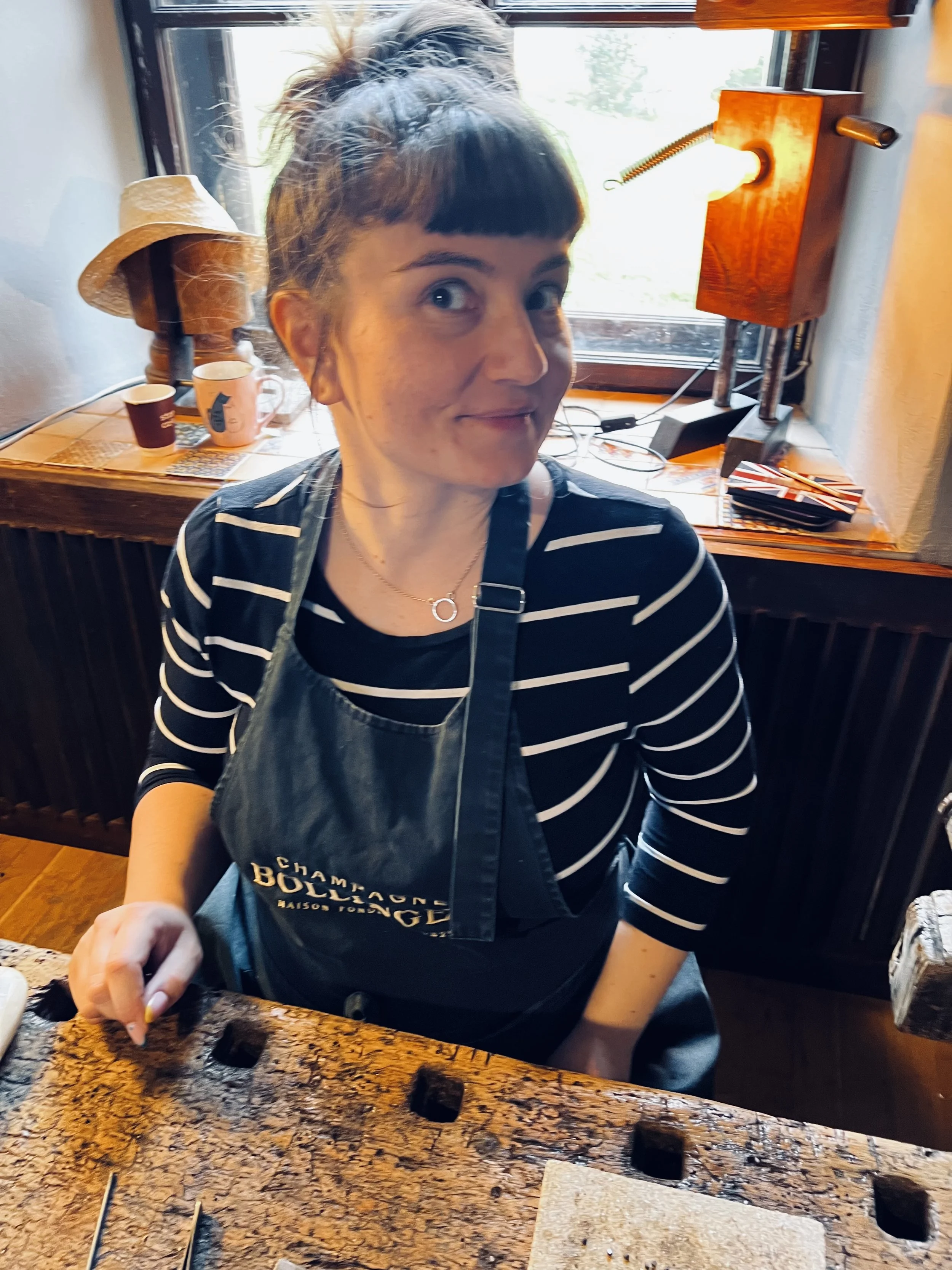 A woman with glasses and a striped shirt is preparing food in a kitchen. She is wearing a black apron with gold lettering. Behind her, there is a window, a lamp, and some cups, one with a blue house design and the other plain white. A hat rests on th