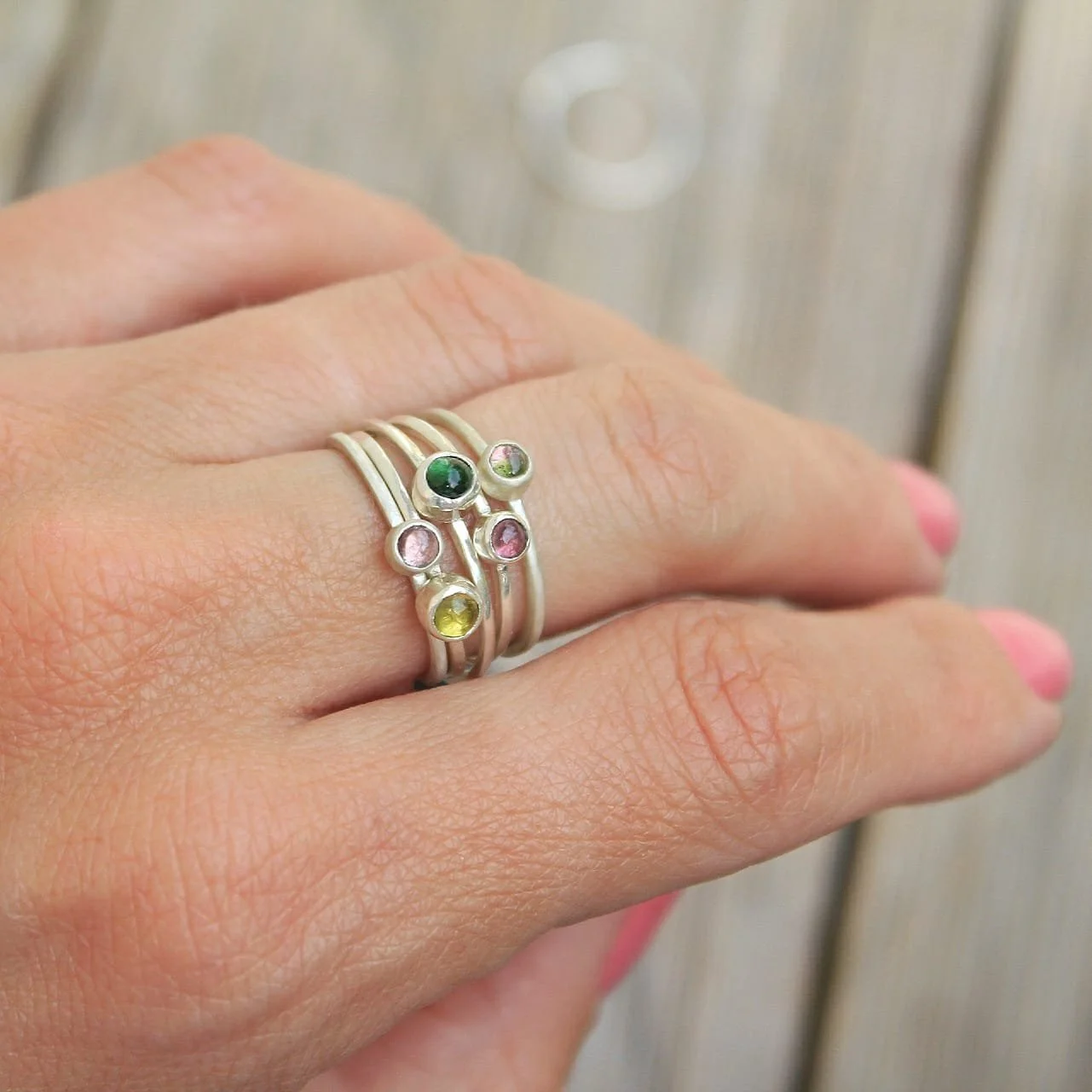 A hand wearing multiple silver rings with colorful gemstones, including green, pink, and yellow stones, with a blurred background of a wooden surface.