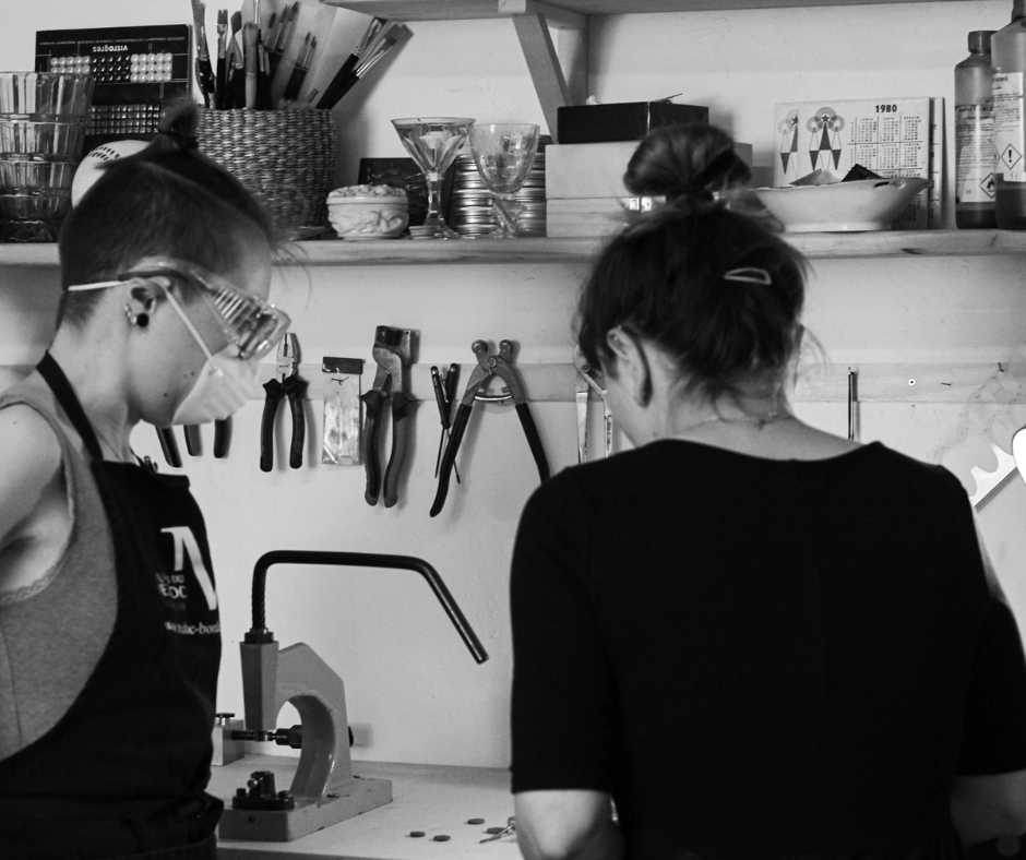 Two women working in a laboratory or workshop, with tools hanging on the wall and various glassware and supplies on shelves.