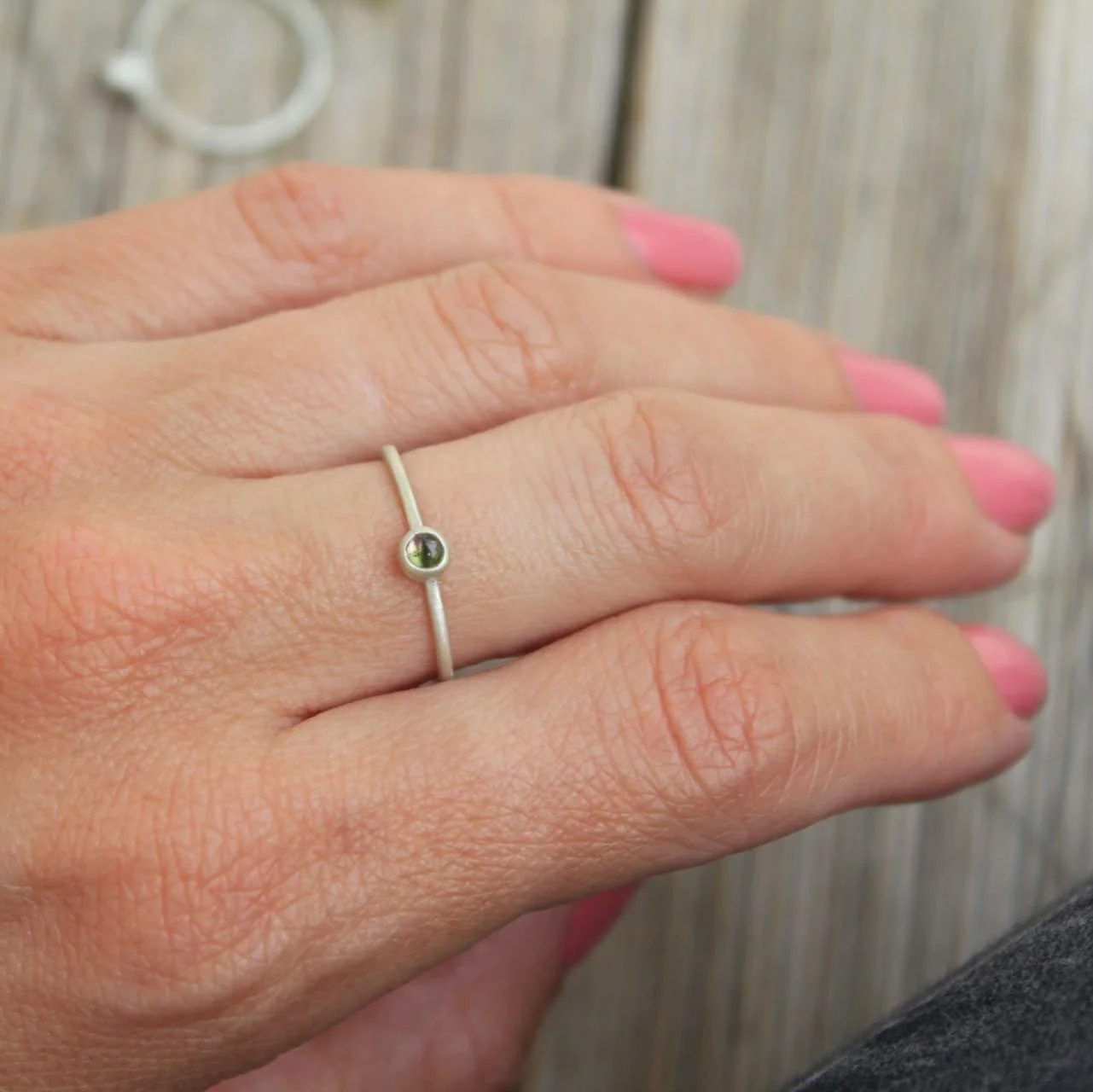 Close-up of a hand with a silver ring on the middle finger. The ring features a small, round, green gemstone. The hand has pink nail polish and is resting on a wooden surface.
