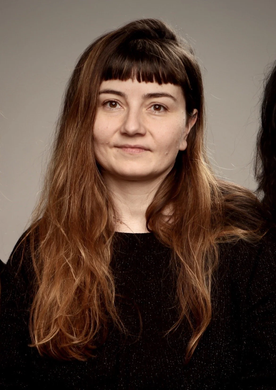A woman with long, wavy brown hair with bangs, wearing a black top, looking at the camera with a neutral expression.