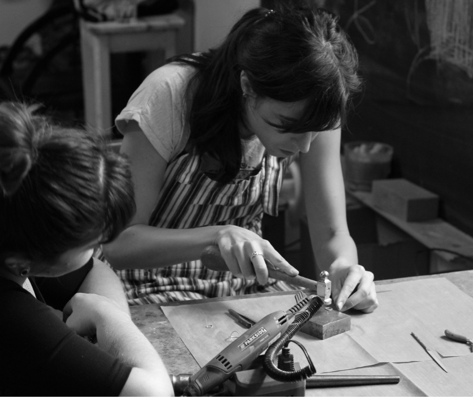 Two women working together at a woodworking bench, one using a hammer and chisel on a piece of wood, while the other observes. They are focused on the craft project.