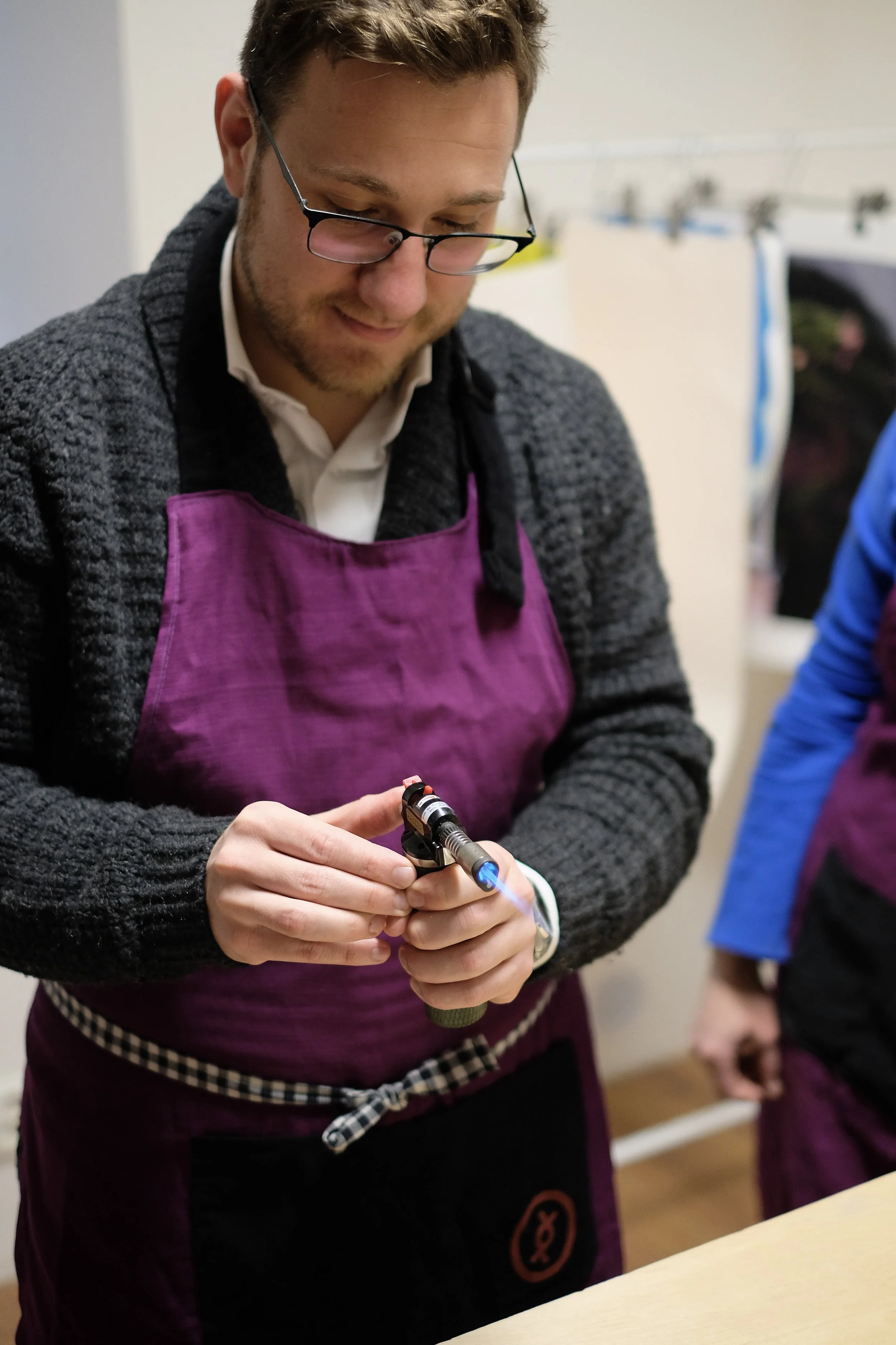 A man with glasses wearing a purple apron and a gray sweater holding a blowtorch under a flame.