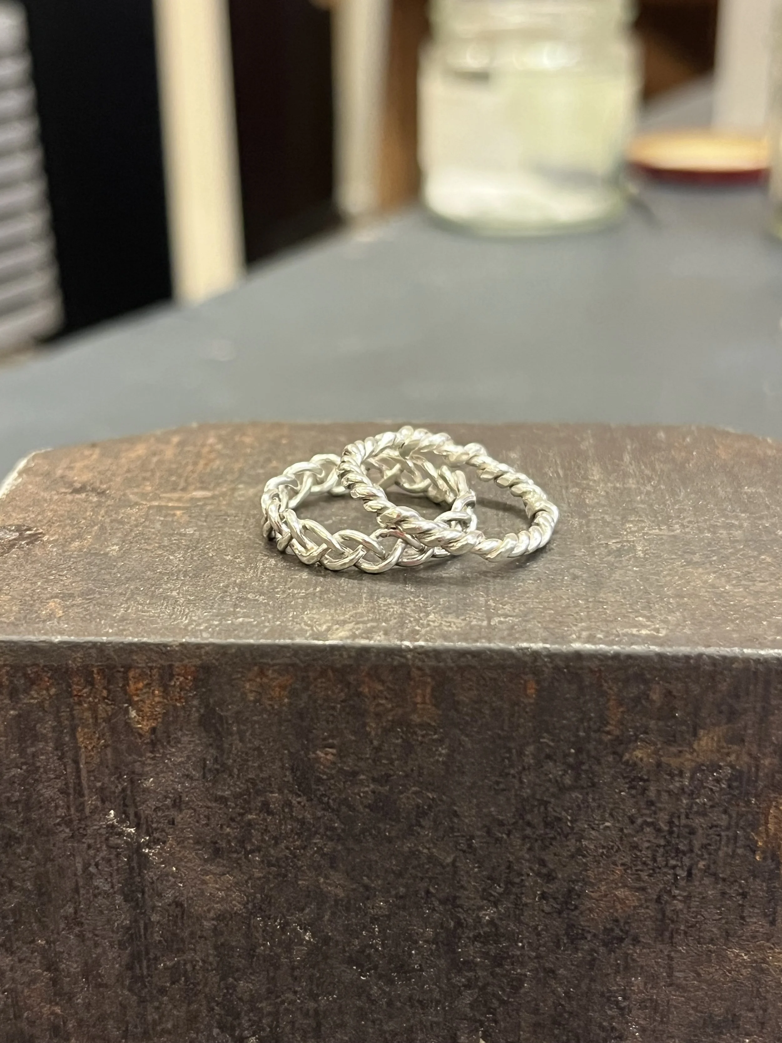 A pair of silver braided rings resting on a textured surface with a mirror-like shine.