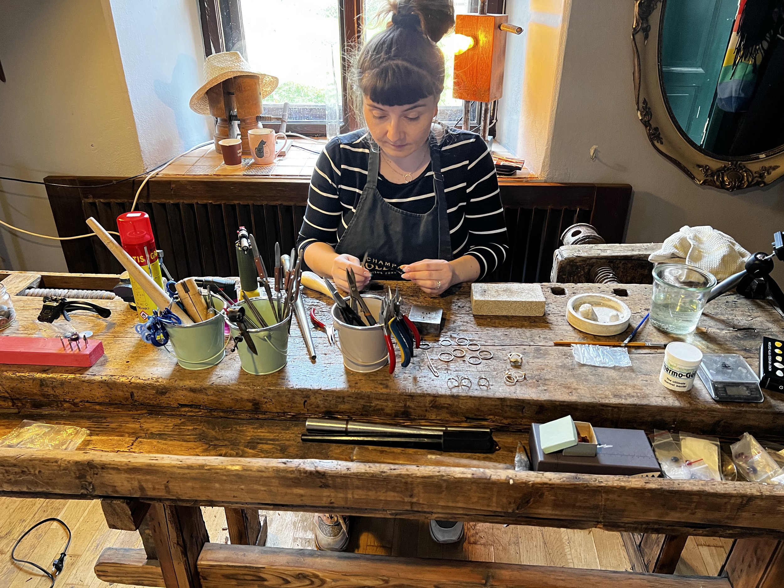 Woman working at a woodworking or jewelry crafting bench with various tools, a glass of water, and a mirror in the background.