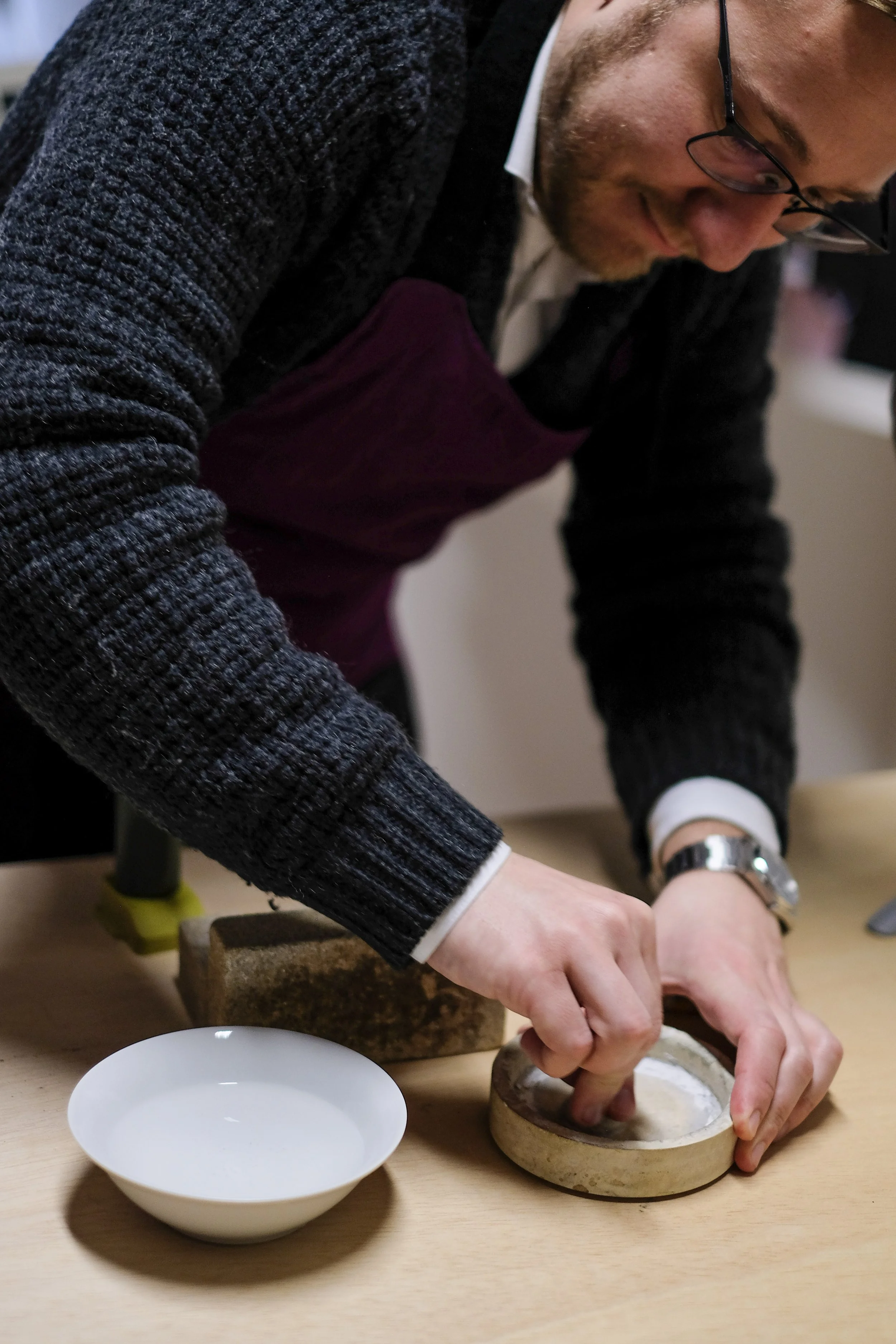 Close-up of a person wearing a wristwatch, leaning over a table and working with their hands to cut or carve a piece of material inside a round container with white powder. A white bowl is on the table in the foreground.