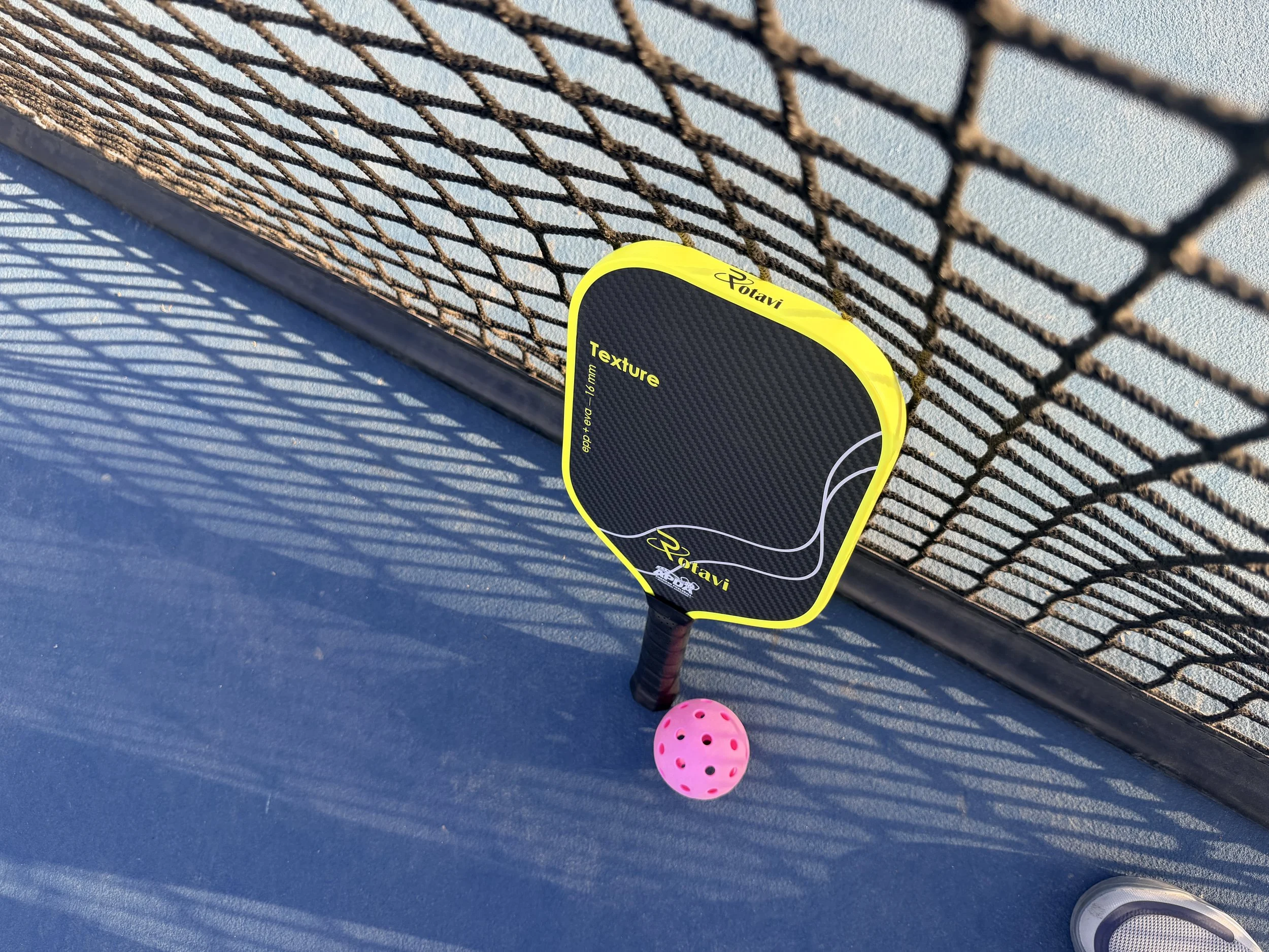 Rotavi Pickleball paddle with yellow and black colors resting against a net, next to a pink pickleball on a blue outdoor court with shadows of the fence cast on the ground.