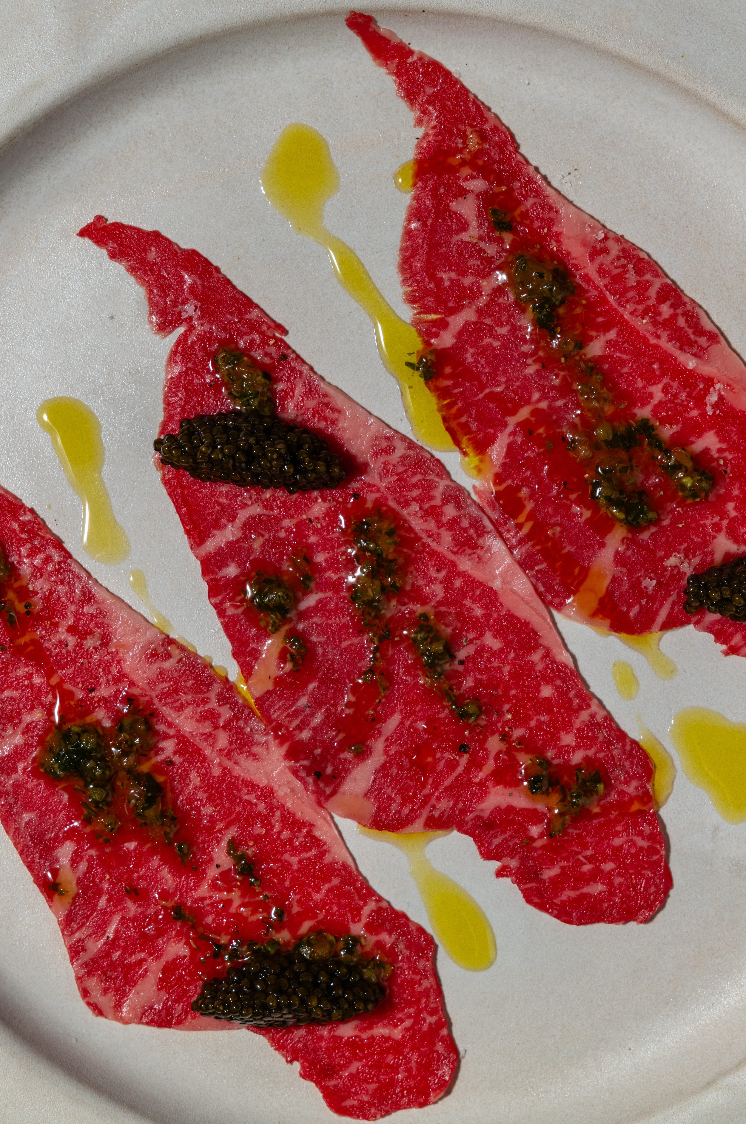 Slices of raw beef steak topped with black caviar and green sauce, drizzled with olive oil on a white plate.