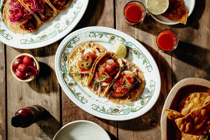 Three tacos filled with meat, salsa, and sliced jalapeños on a decorative oval plate with lemon wedges, with two small glasses of red sauce, a bowl of radishes, a drink in a glass bottle, and additional dishes of fried food on a rustic wooden table.