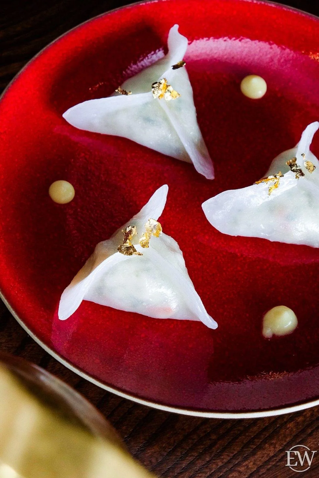 Handmade dumplings garnished with edible gold leaf, served on a red plate with small dollops of white sauce or cream.