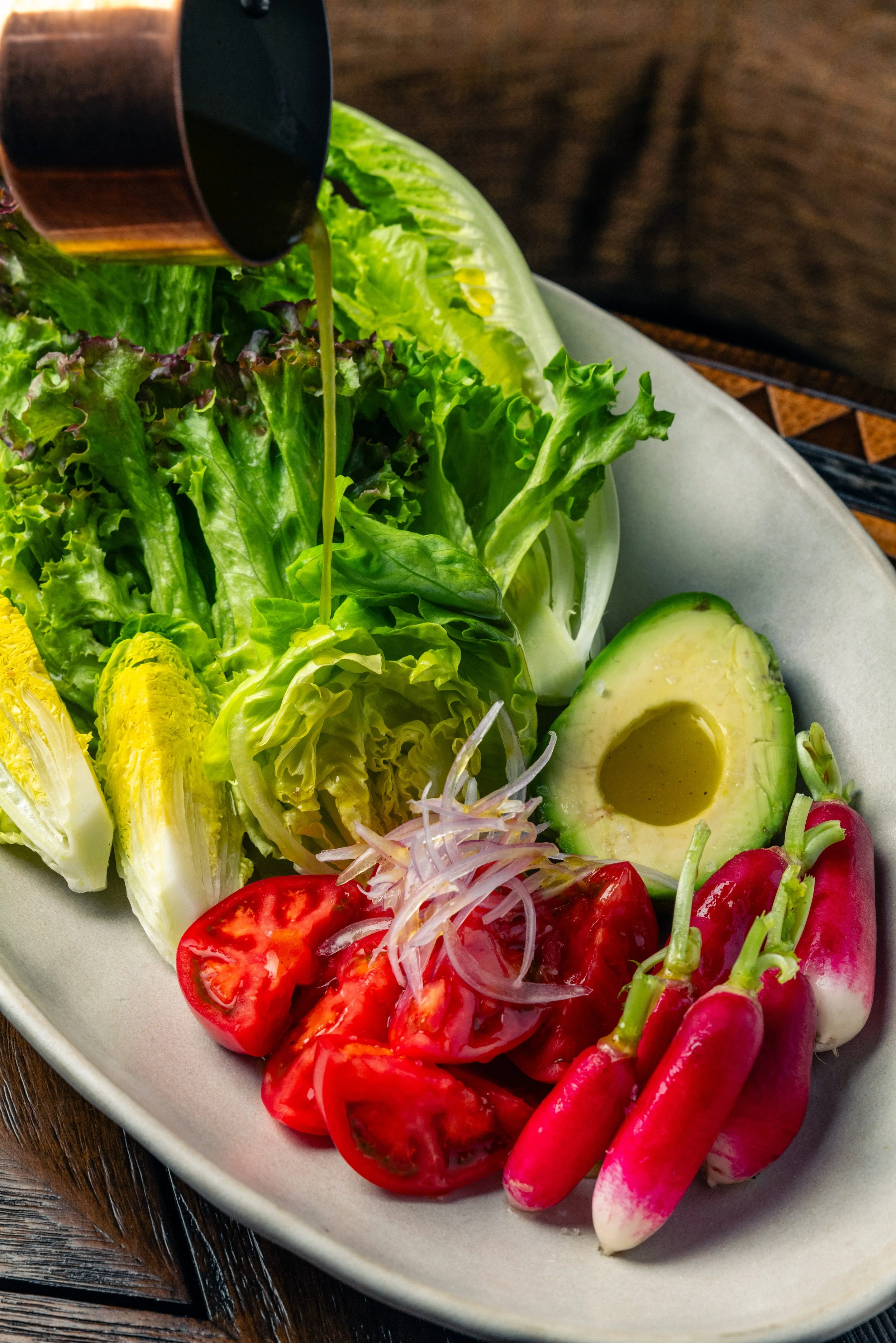 Fresh vegetables on a white plate including lettuce, tomatoes, radishes, cucumber, and avocado, with a dark beverage being poured over the greens.
