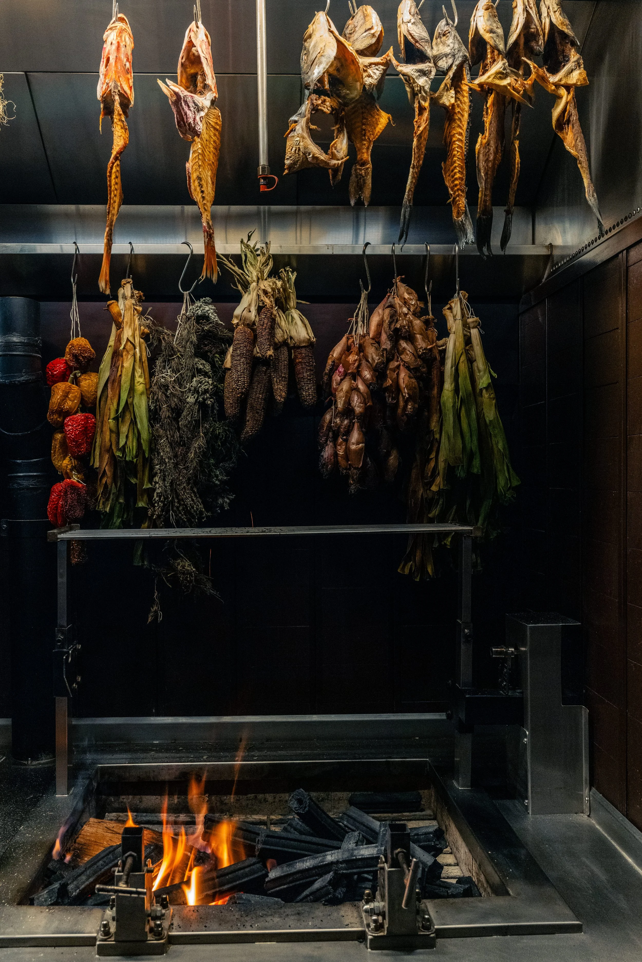 Dried fish, herbs, and vegetables hanging above a wood fire in a kitchen.