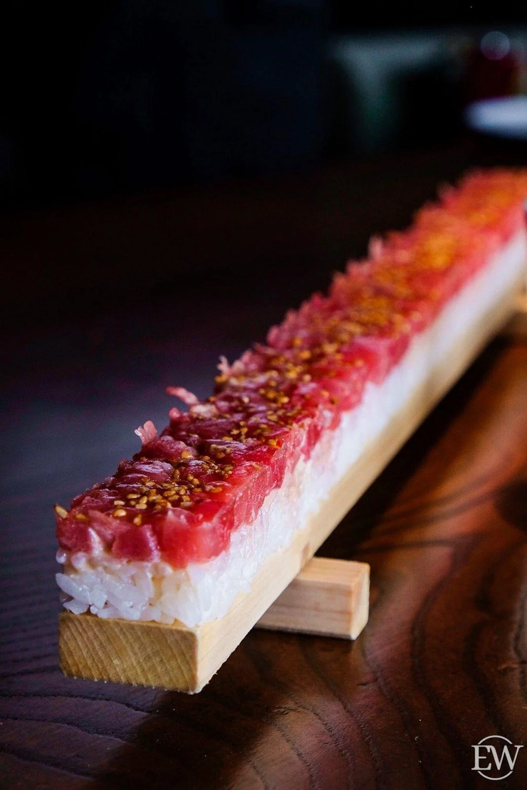 Close-up of a long sushi roll with raw fish and topped with seeds, placed on a wooden serving board.