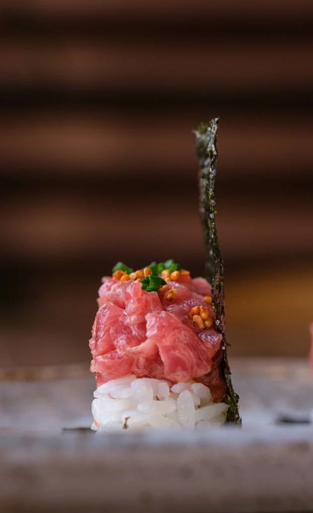 Close-up of a piece of sushi with diced raw tuna, topped with orange roe and chopped green onions, garnished with a vertical strip of seaweed, on a wooden surface.