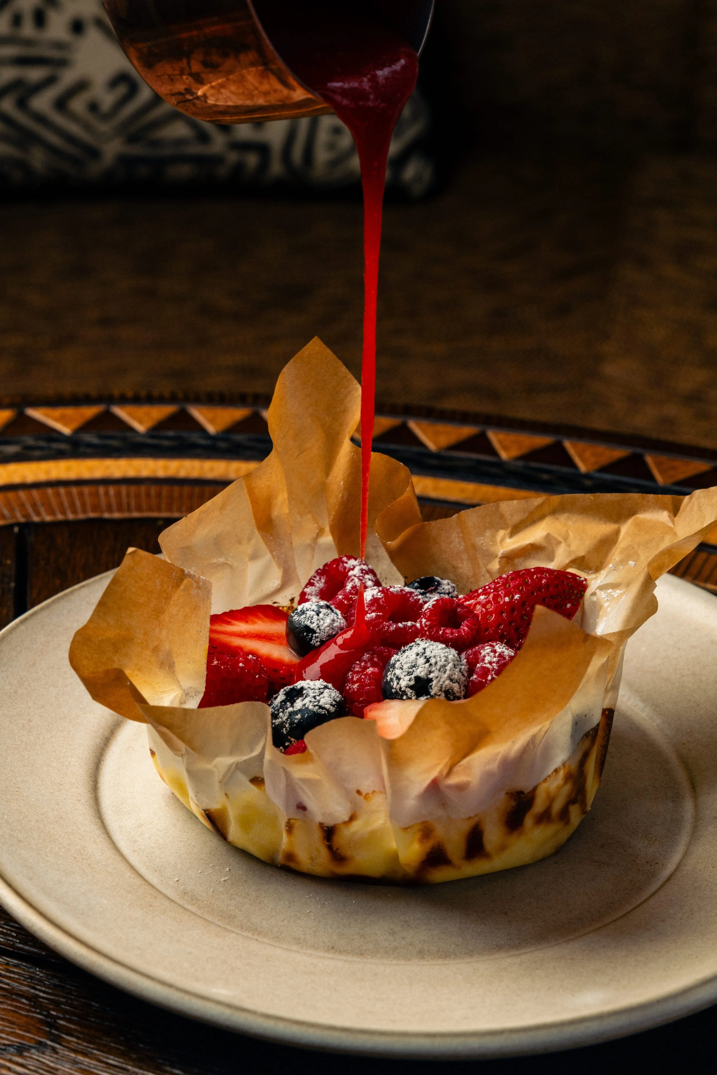 A dessert bowl with mixed berries, including strawberries, blueberries, and raspberries, topped with powdered sugar, with red sauce being poured over it.