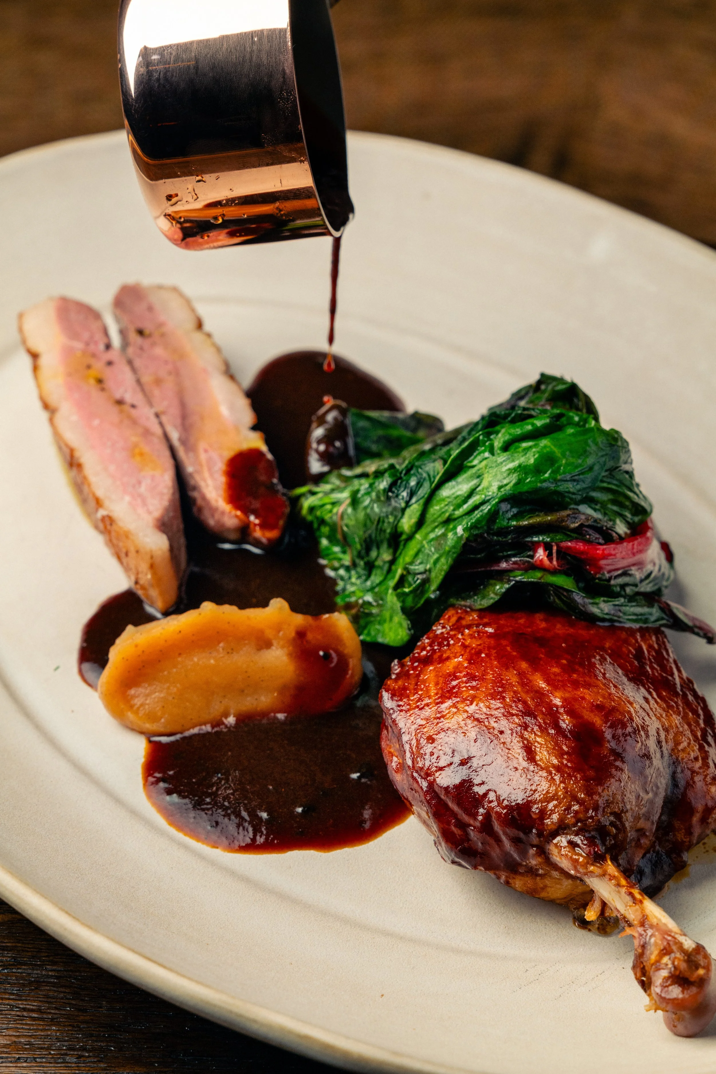 A plated gourmet meal with sliced duck breast, a stuffed potato, a green leafy vegetable, and a roasted chicken leg with sauce being poured over it.