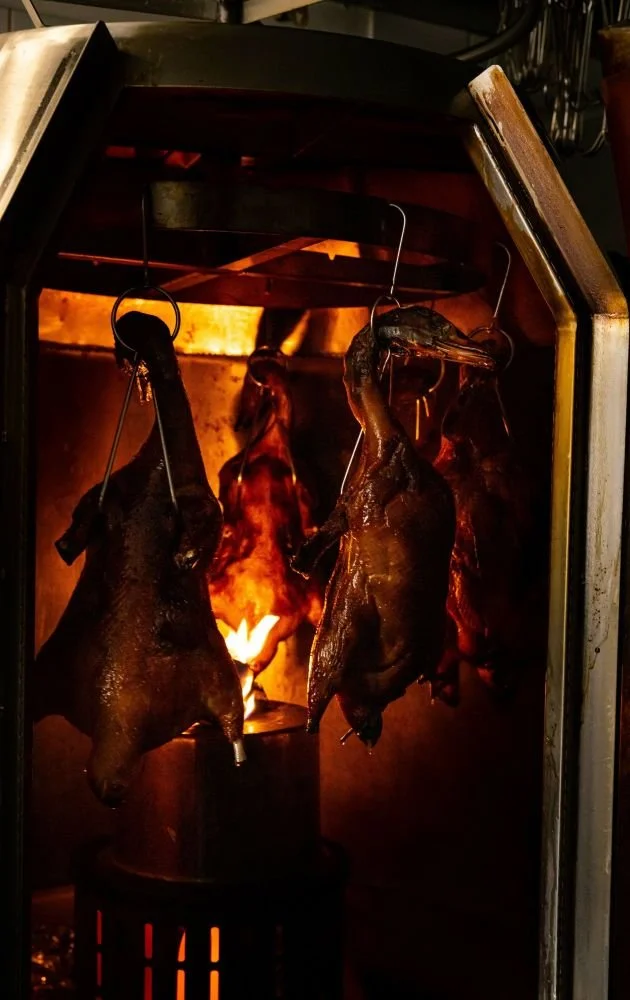 Three ducks roasting on a spit over an open flame inside a rotisserie oven.