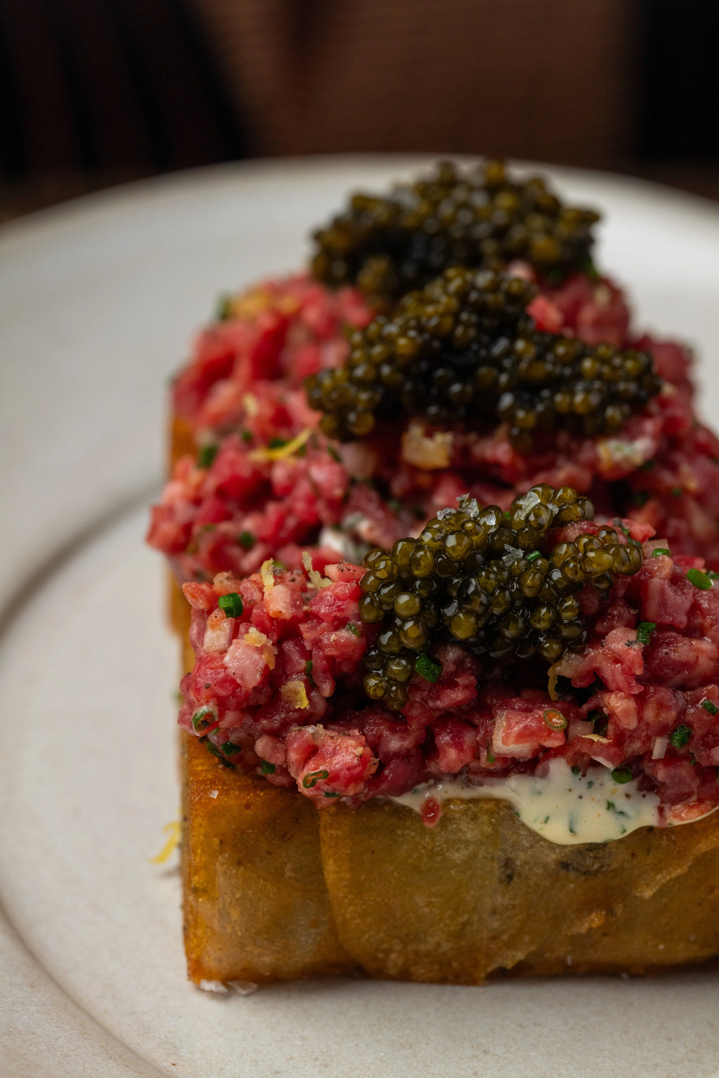 Close-up of an appetizer featuring a crispy square base topped with a mixture of minced raw meat, garnished with small green herbs, and topped with black caviar on a white plate.