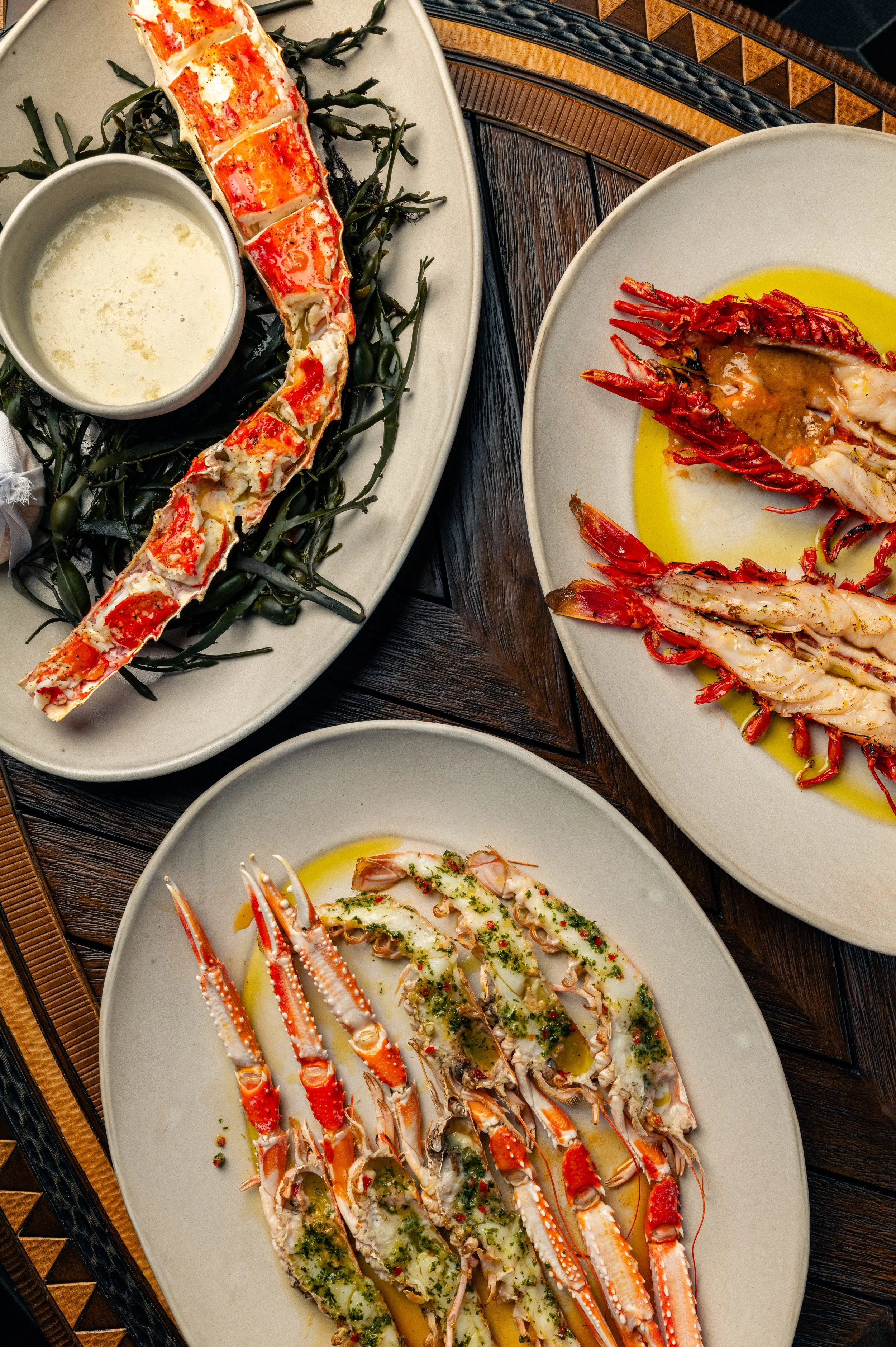 Three plates of shellfish prepared in different styles, with sauces and herbs, on a wooden table.