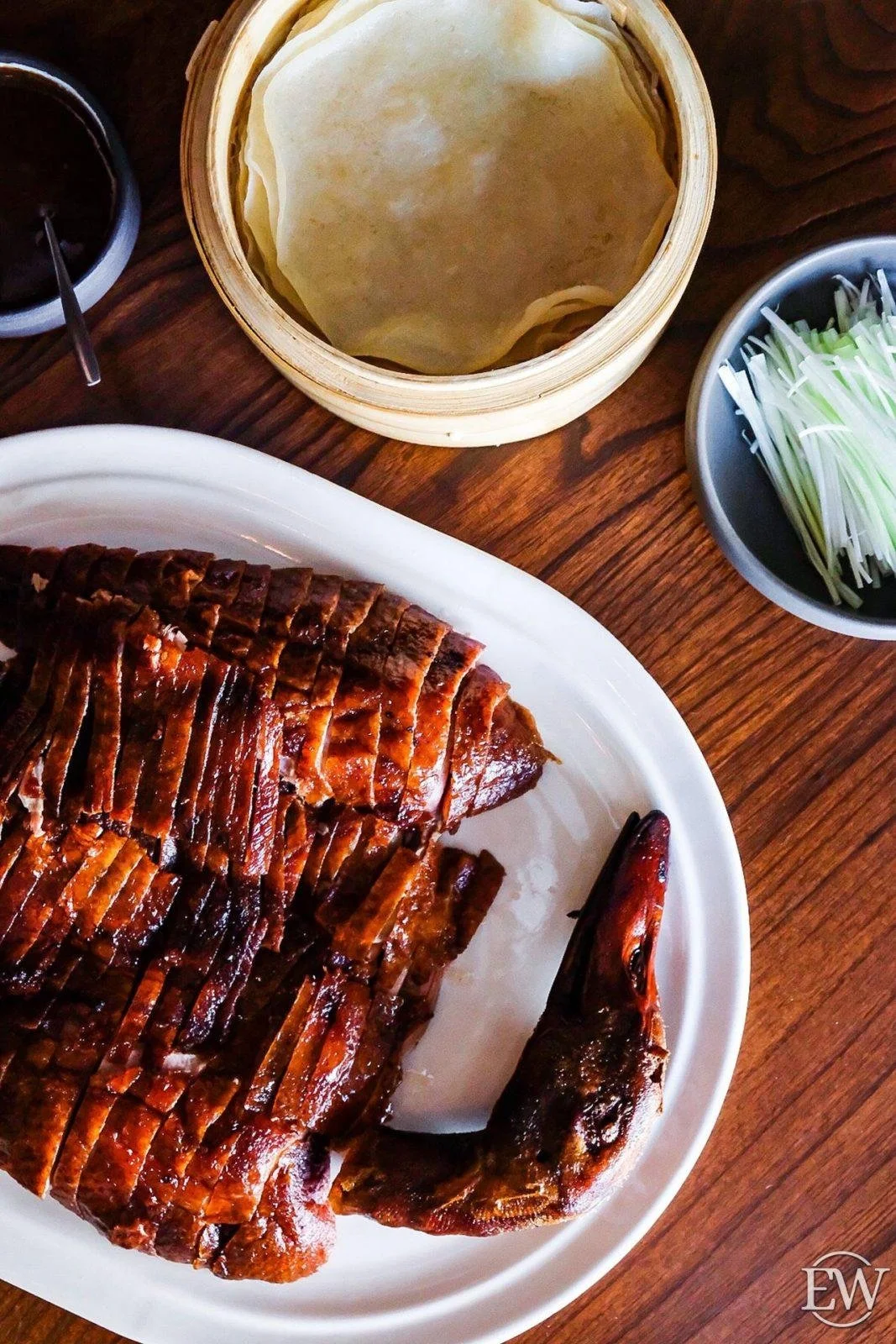 Sliced roasted duck on a white plate with a duck head at the corner, along with a bamboo steamer with dumpling wrappers, a bowl of sliced green onions, and a small bowl of dipping sauce on a wooden table.