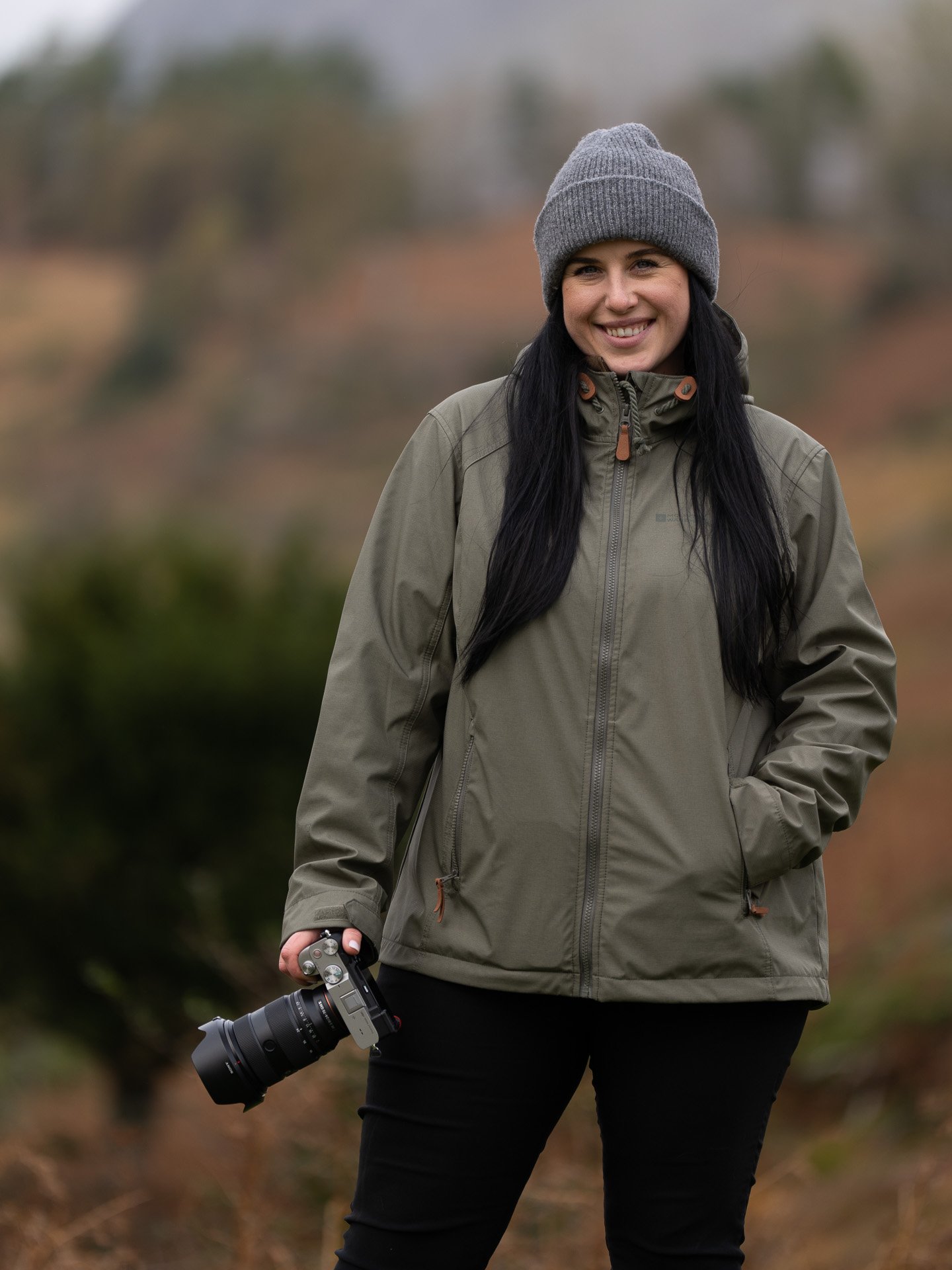 Woman outdoors wearing a grey beanie and a green jacket, holding a camera, smiling, with a blurred natural landscape background. East Yorkshire photographer, product photography, pet photography, nature photography