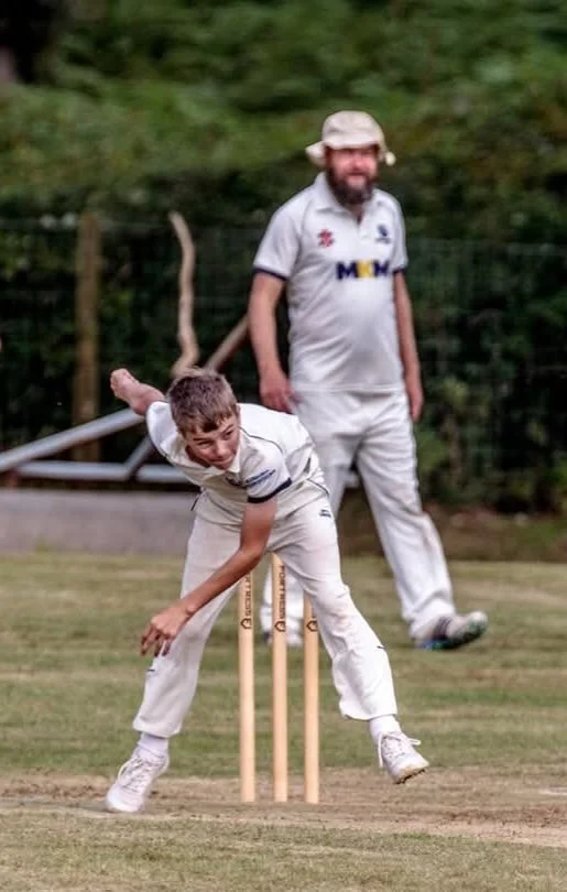 A young boy bowls in a cricket game on a grassy field, while an adult man stands in the background watching. The boy is mid-action, with a focused expression, dressed in white cricket attire, and the man wears a white hat and shirt.