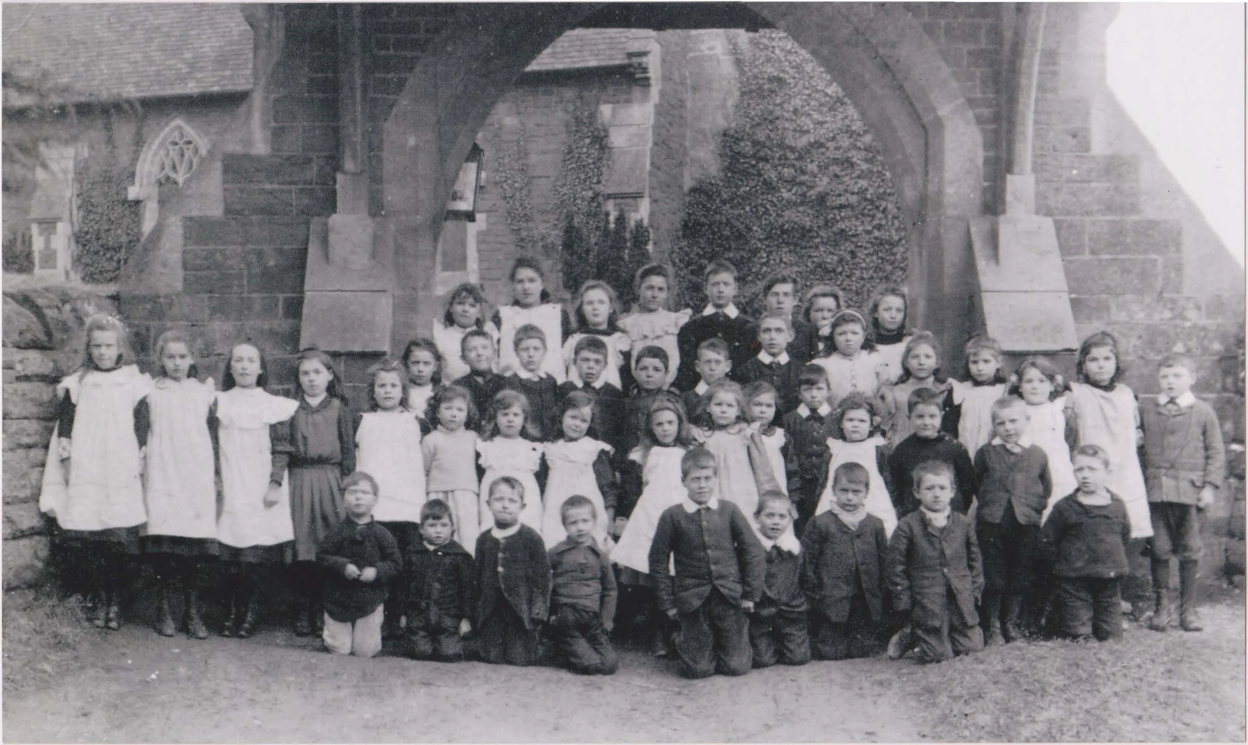 Black and white photo of children from Oxenhall School. The children are arranged in multiple rows, some standing and some kneeling or sitting, dressed in old-fashioned clothing.
