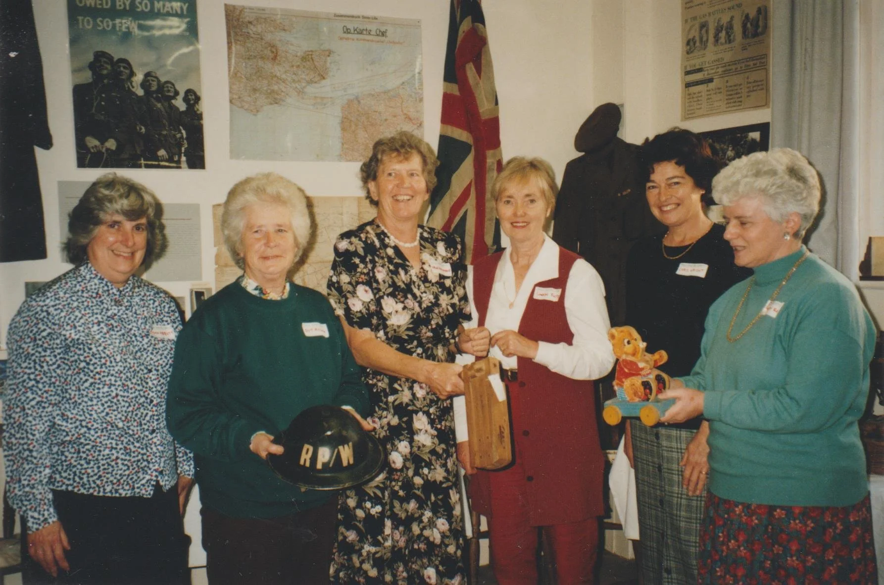 A group of seven women standing indoors, some holding items like a black helmet with 'RPW' letters and a teddy bear on a children's wagon. The background features racial posters, a map, and a British flag.