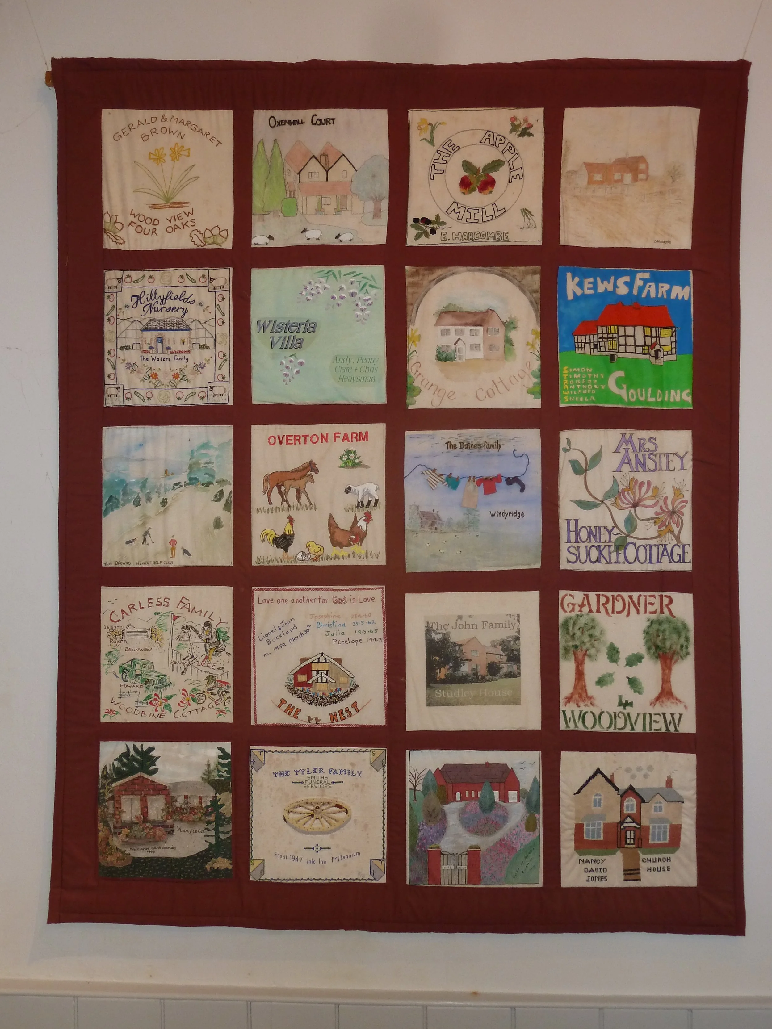 A quilt display featuring colorful embroidered and painted patches of various family names, houses, farm scenes, and decorative motifs.