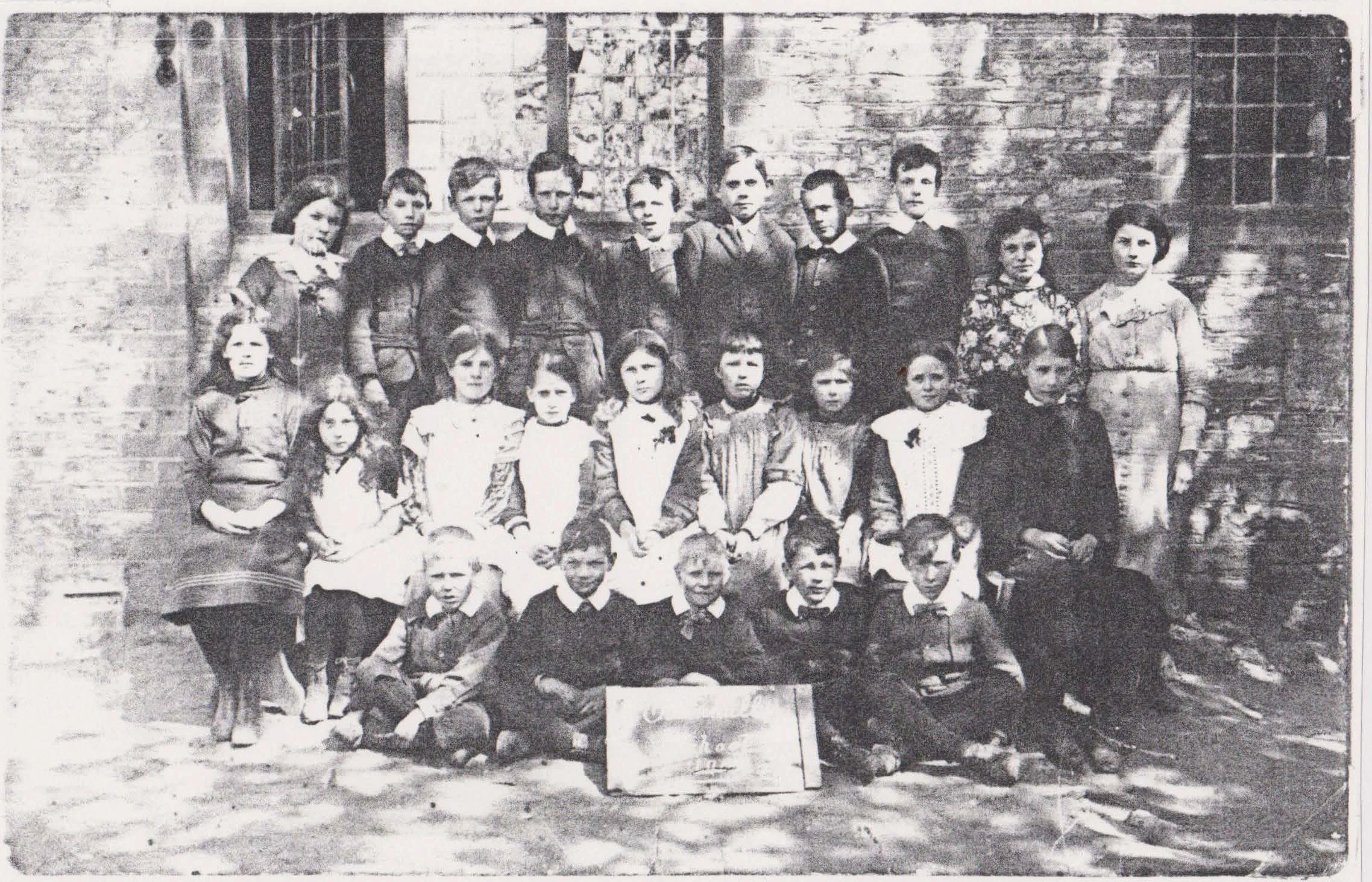 Black and white photograph of a group of children from Oxenhall School and their teacher posing outside a brick building. The children are arranged in three rows, some sitting, some standing, with a chalkboard or sign in front.