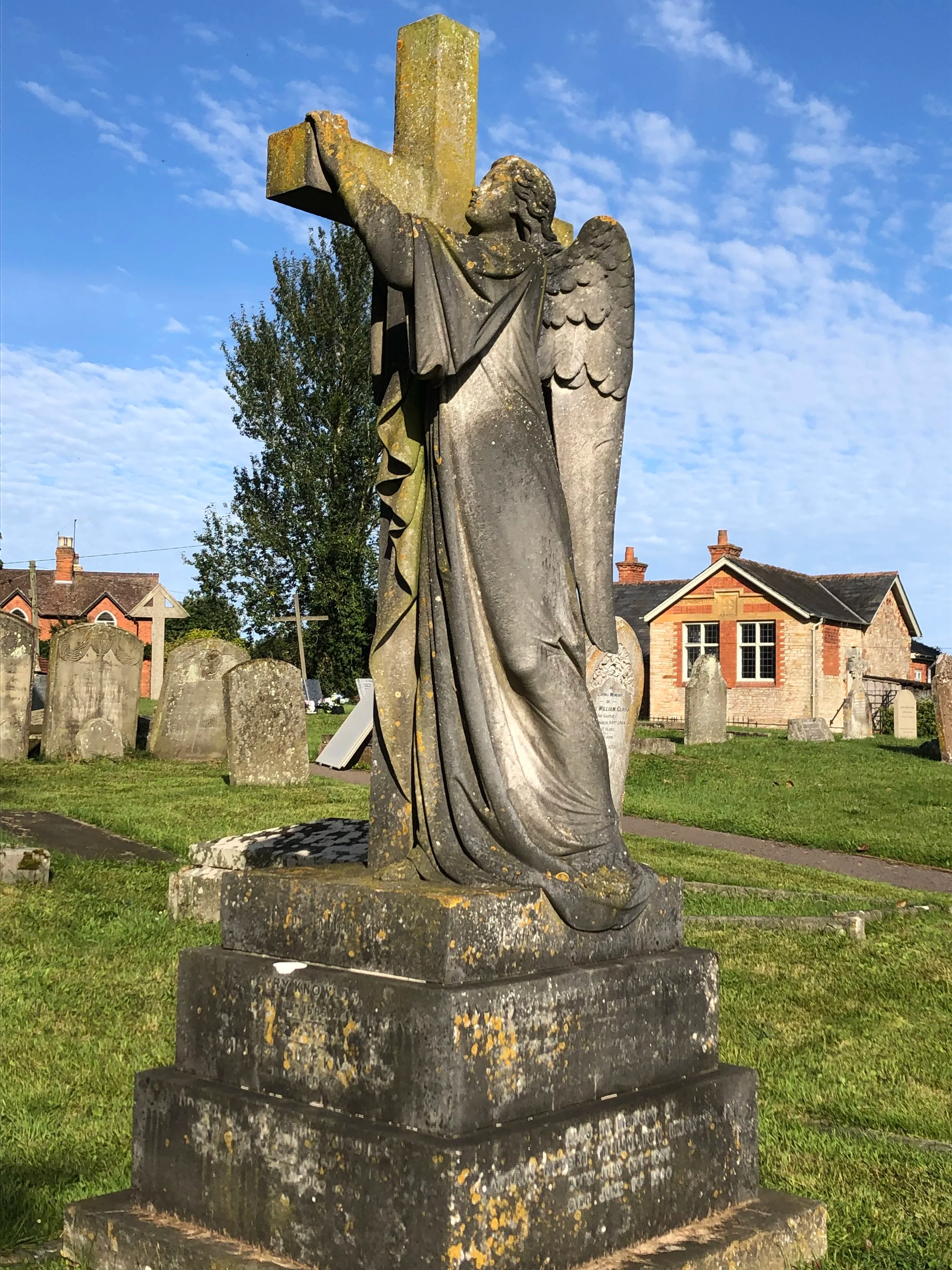 A stone statue of an angel with wings, holding a cross, in a graveyard with tombstones and a building in the background under a partly cloudy sky.