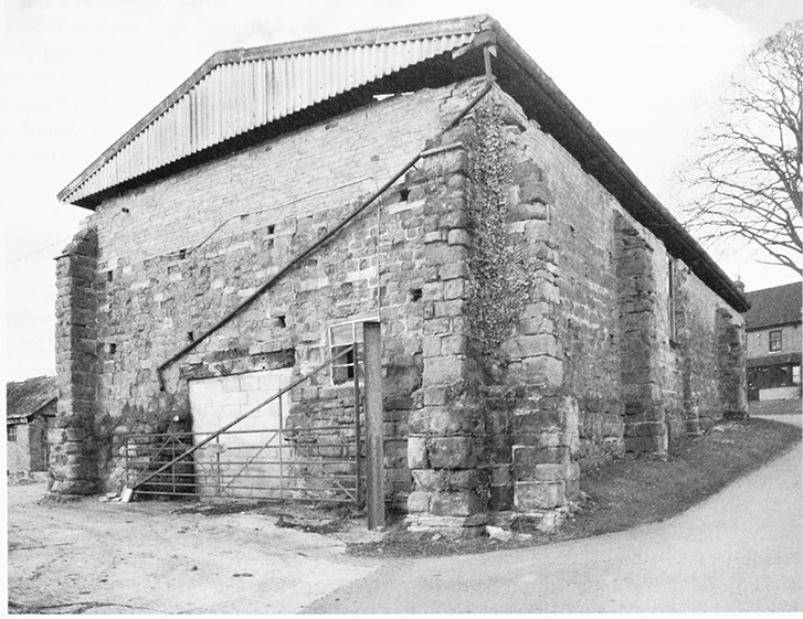 Black and white photo of an old stone barn with a corrugated metal roof, surrounded by a dirt ground and a few trees in the background.