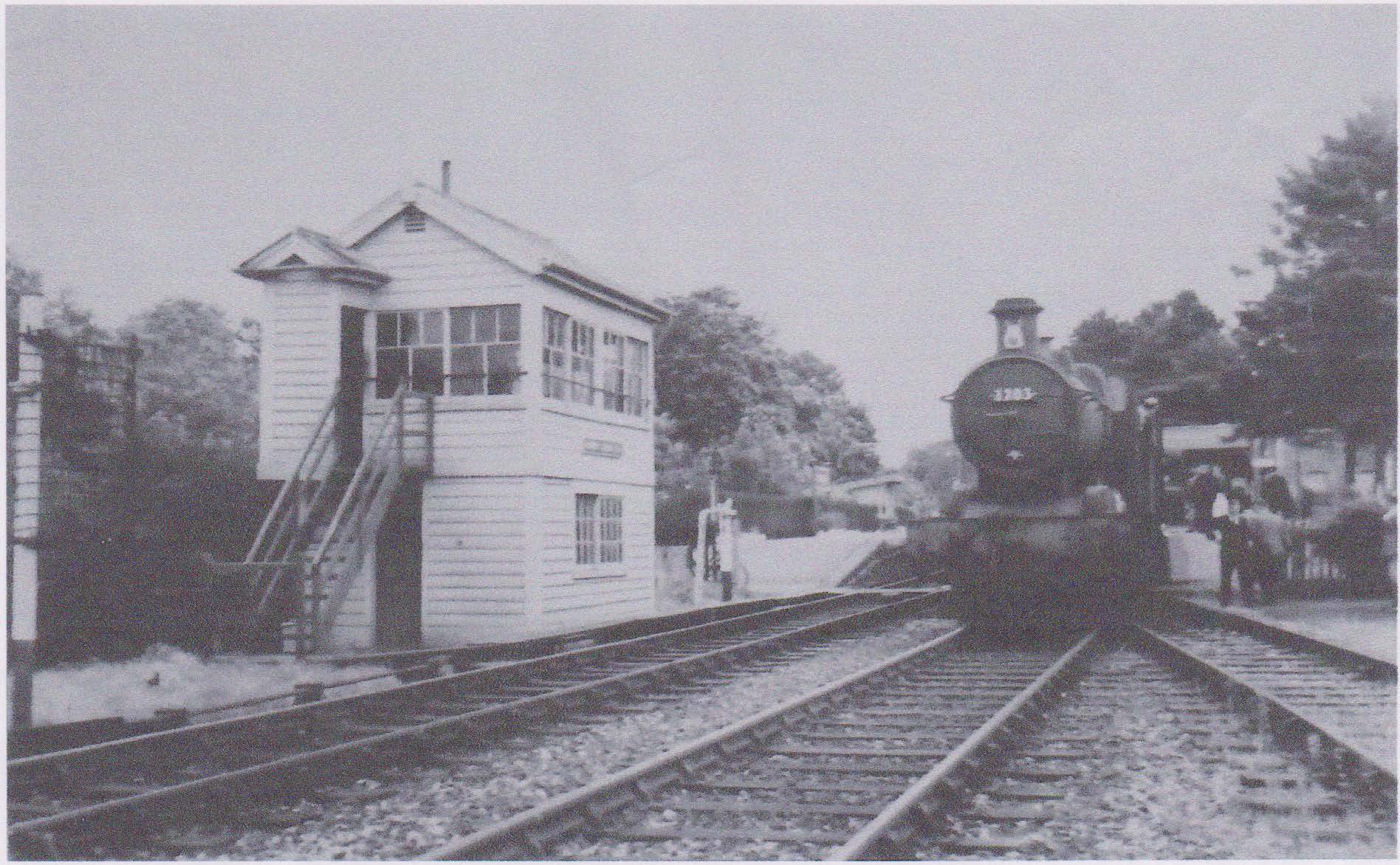 A vintage black and white photograph of a train station with a locomotive approaching. There is a small building with stairs on the left and trees in the background.