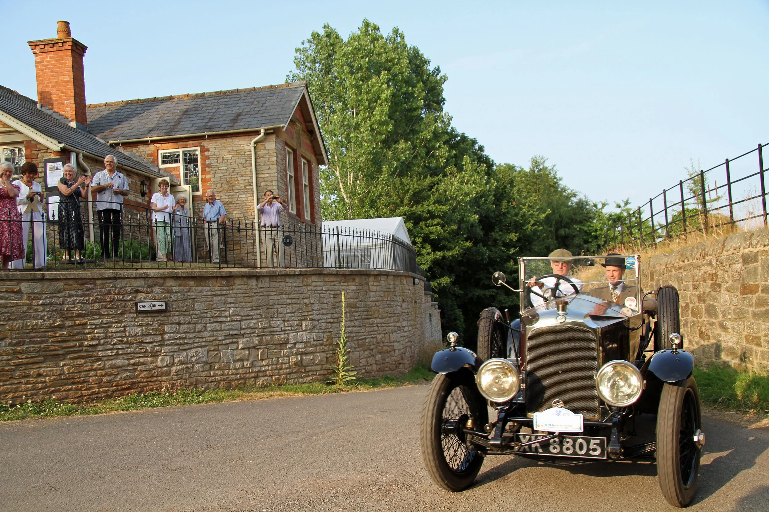 Vintage car driving past a group of people standing on an elevated porch of a stone building with a brick chimney, during daylight with trees in the background.  Outside St. Anne's church Oxenhall.