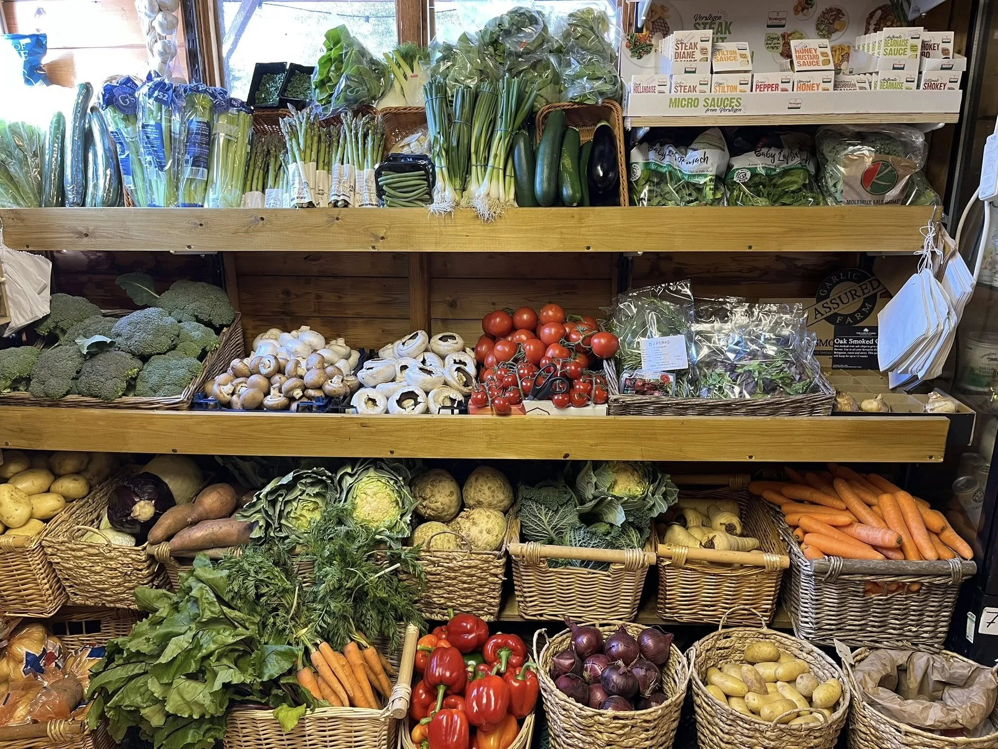 Display of fresh vegetables on wooden shelves and in baskets, including broccoli, mushrooms, tomatoes, green leafy vegetables, carrots, potatoes, onions, and eggplants.
