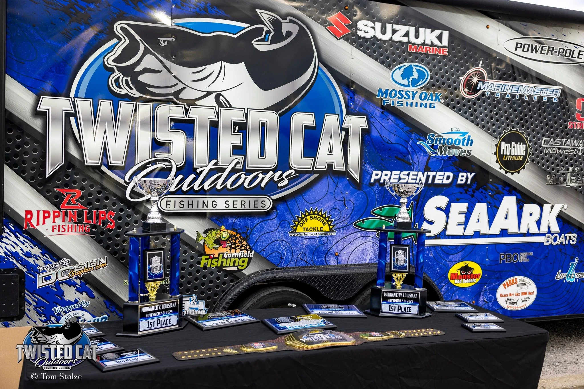 Trophies and awards from the Twisted Cat Outdoors fishing series displayed on a table, with the Twisted Cat Outdoors trailer in the background showing sponsors, including Suzuki Marine, Mossy Oak Fishing, and SeaArk Boats.