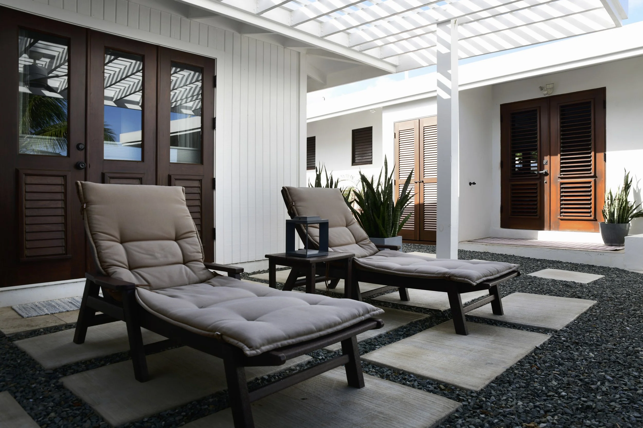 Outdoor patio with two beige cushioned lounge chairs, side table, black lantern, potted plants, white walls, wooden doors, and a partially covered roof.