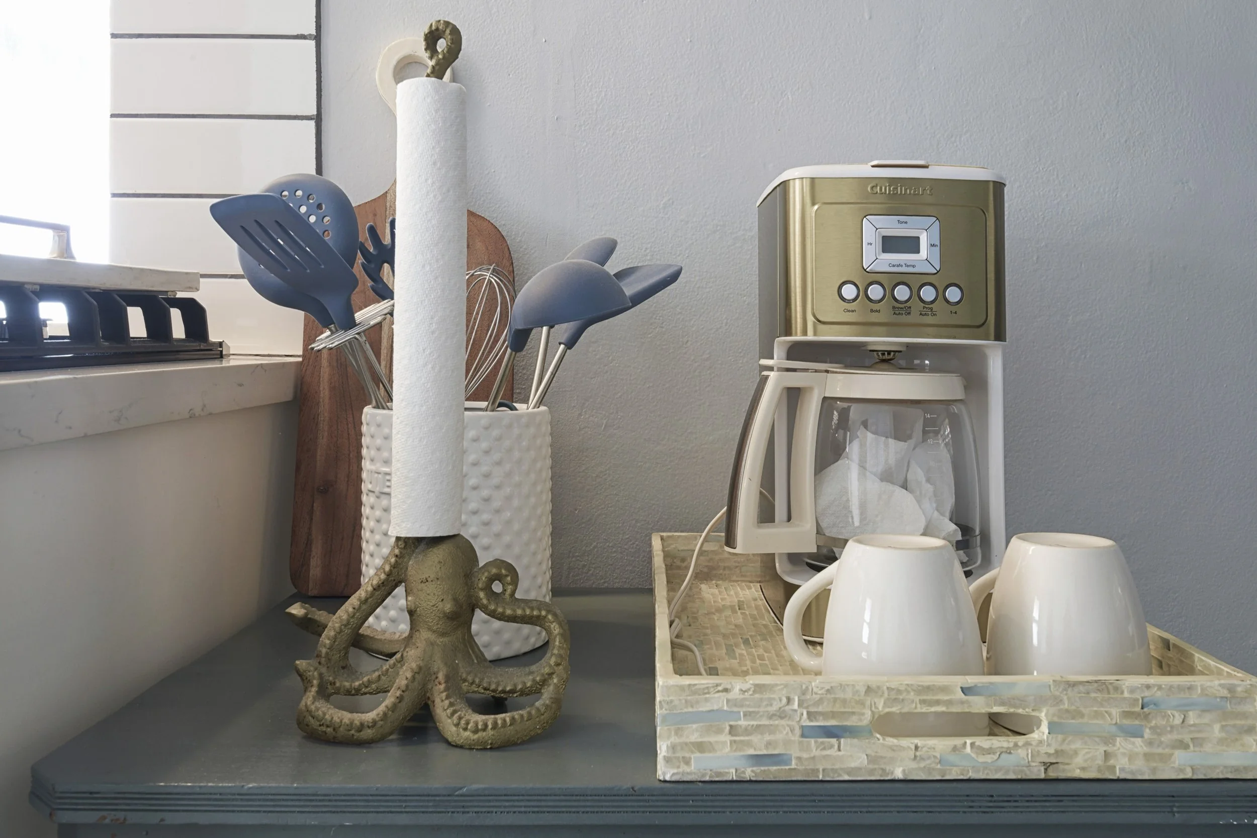 Kitchen countertop with a paper towel holder shaped like an octopus, utensils in a white textured container, a wooden cutting board, and a coffee maker with two cups on a tray.