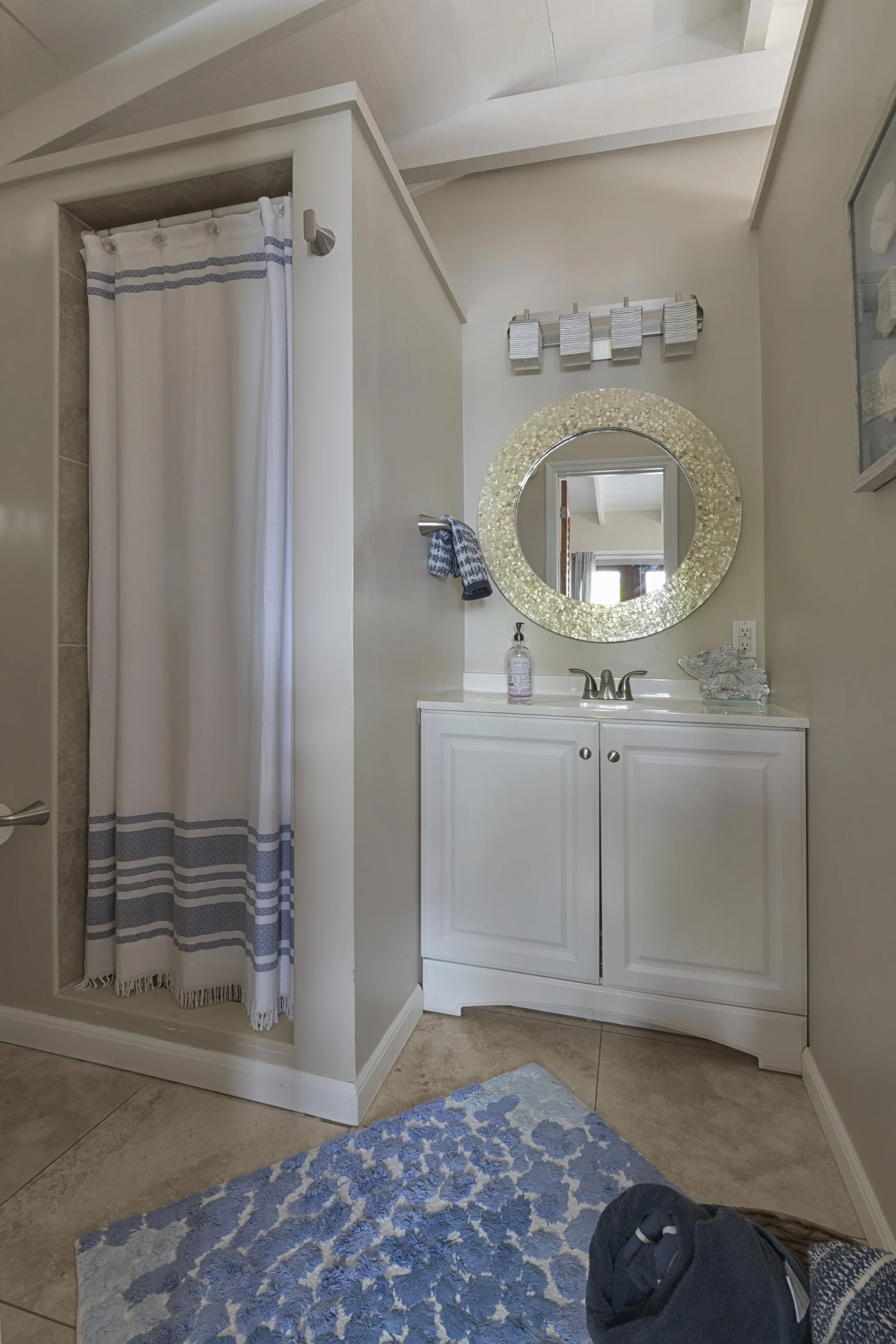 A bathroom with a white vanity, a round mirror with a mosaic gold frame, a small shower area with a curtain, and a blue and white patterned rug.