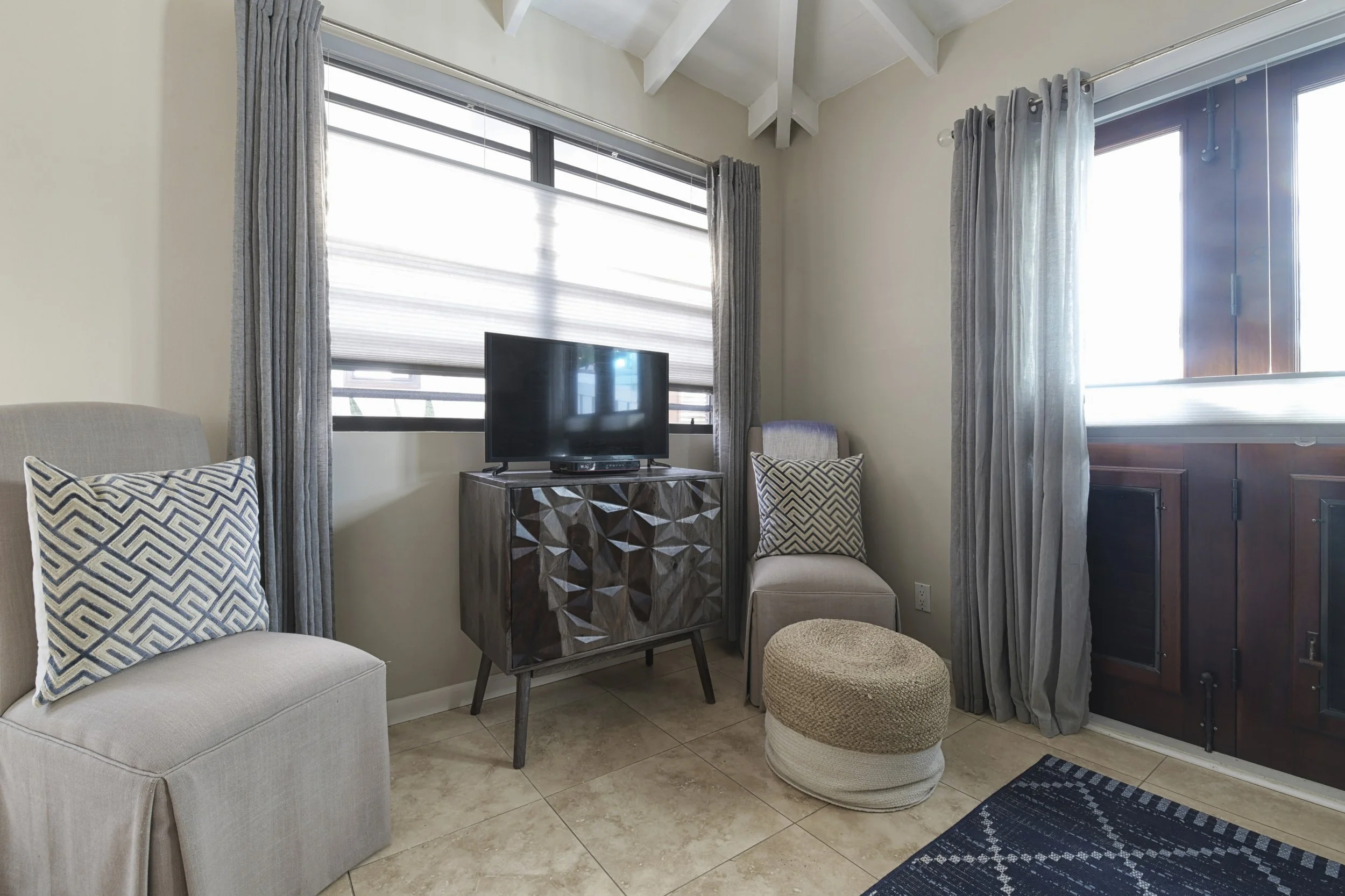 Living room with beige walls, two large windows with grey curtains, a beige armchair with a geometric-patterned pillow, a dark wood TV stand with a small flat-screen TV, a beige armchair with a matching pillow, a round woven ottoman, a patterned rug,