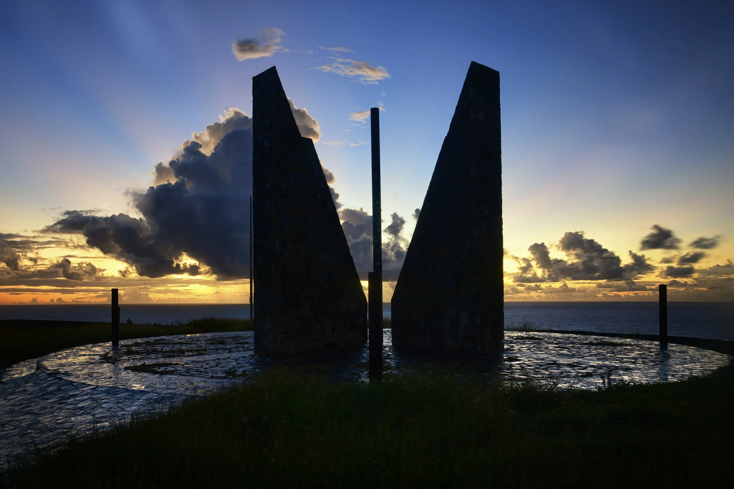 Silhouette of a monument with two large, sloped stone pillars and a central pole, set against a sunset sky over the ocean with scattered clouds.